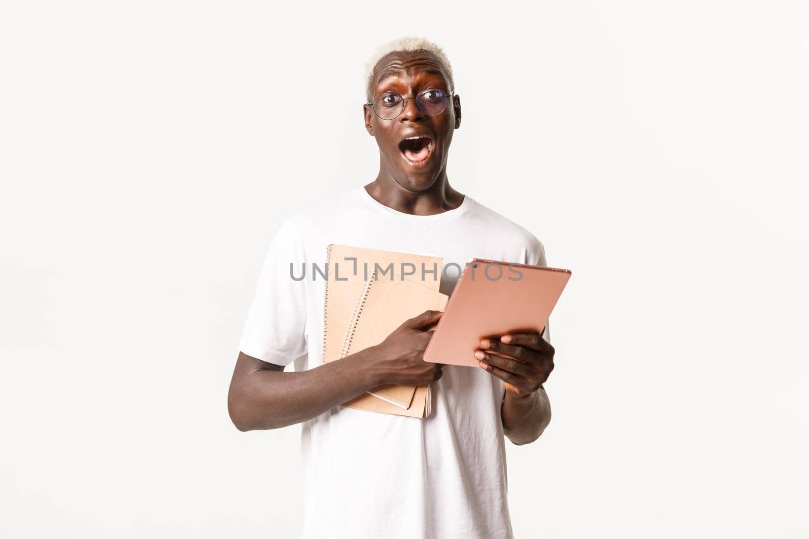 Portrait of amazed, handsome african-american blond male student, wearing glasses, holding notebooks for studying and digital tablet, white background.