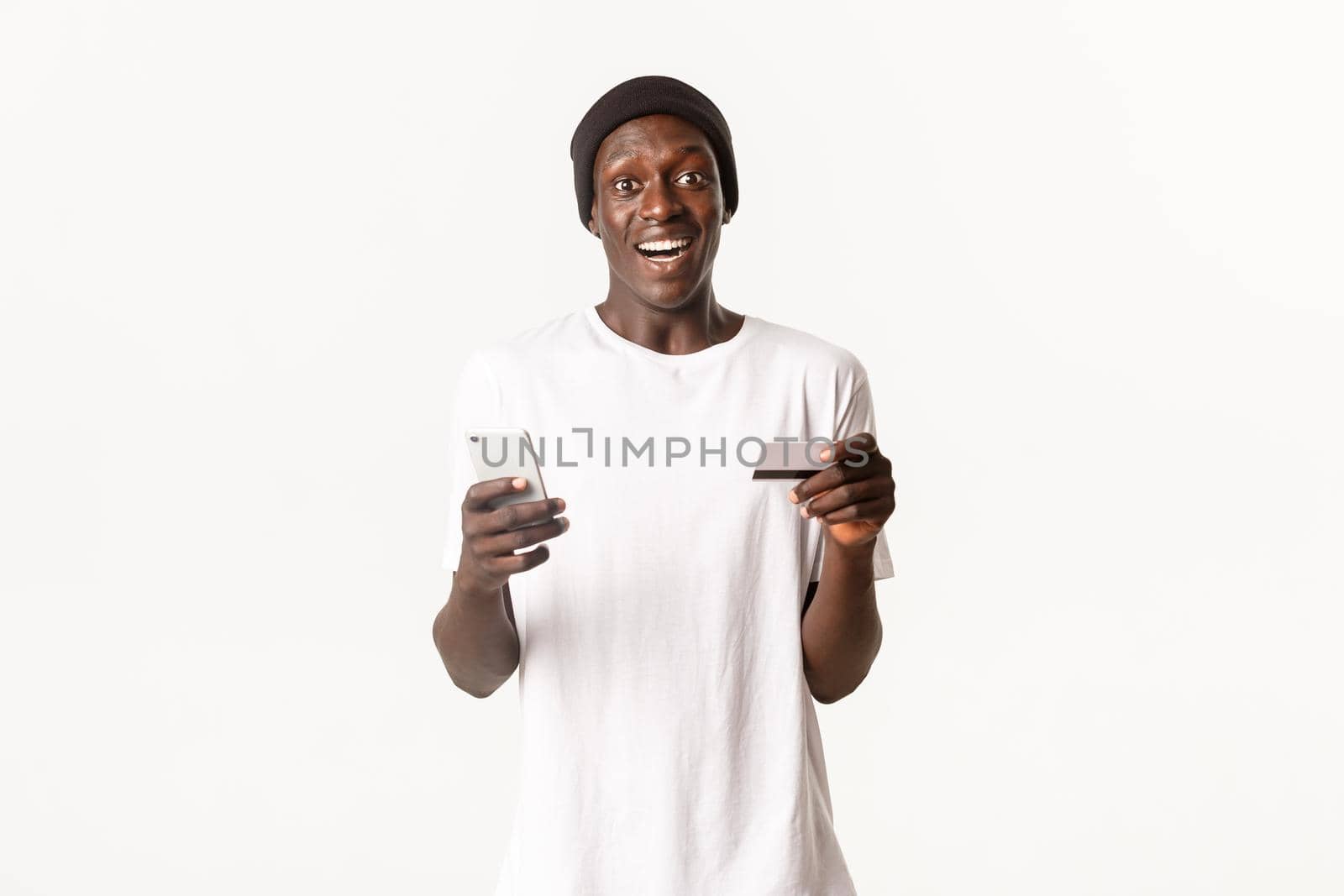 Portrait of excited, happy african-american young male student, holding credit card and smartphone with amused smile, white background by Benzoix