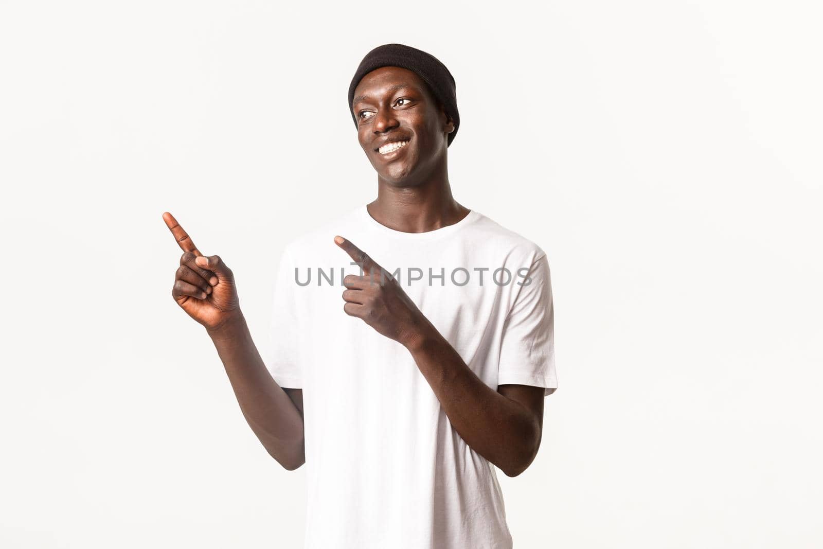 Portrait of happy handsome african-american young man, wearing beanie and t-shirt, smiling pleased and looking, pointing upper left corner, white background.