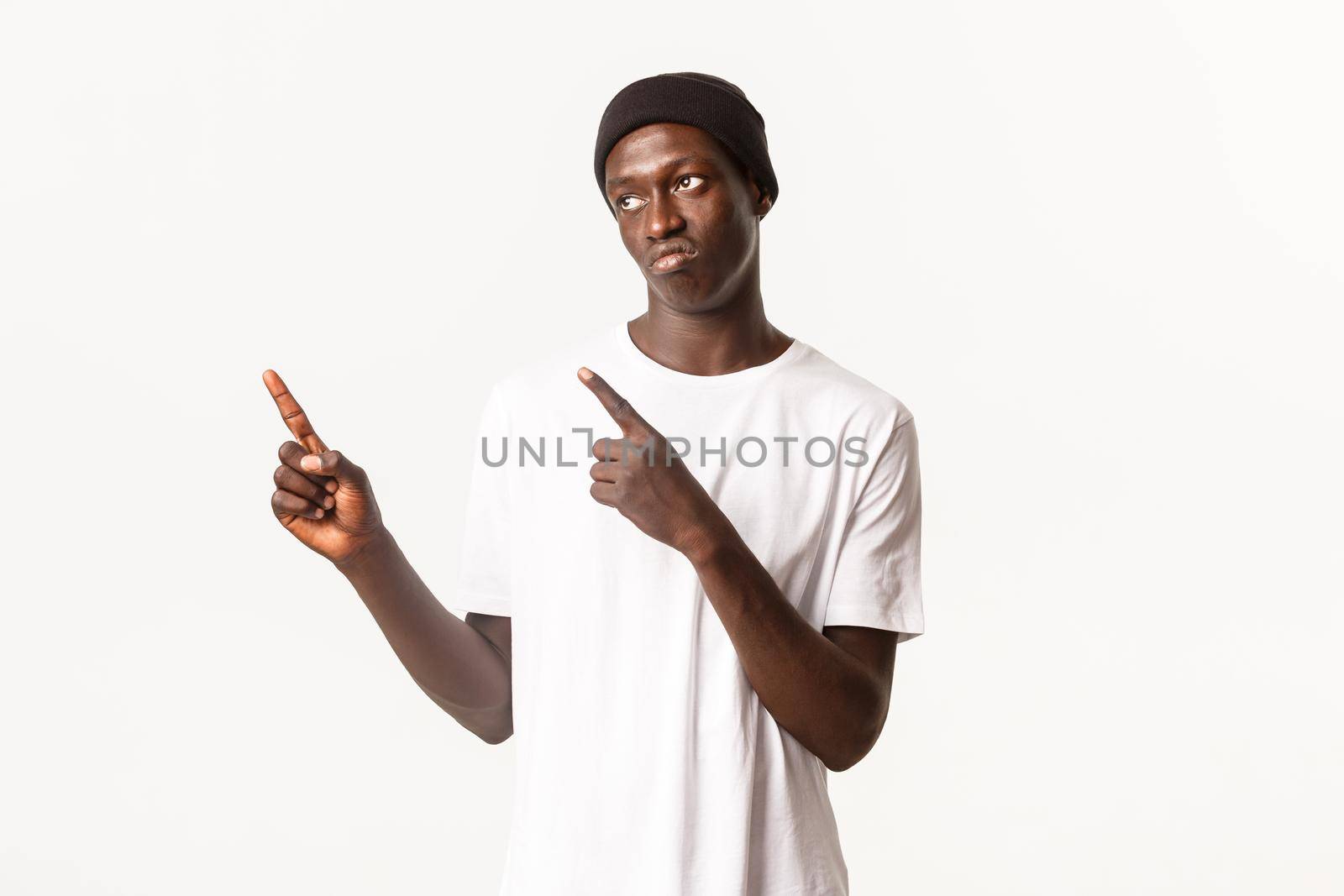Portrait of skeptical or disappointed sassy african-american guy, wearing beanie, pointing fingers upper left corner and sulking, looking upset, white background.