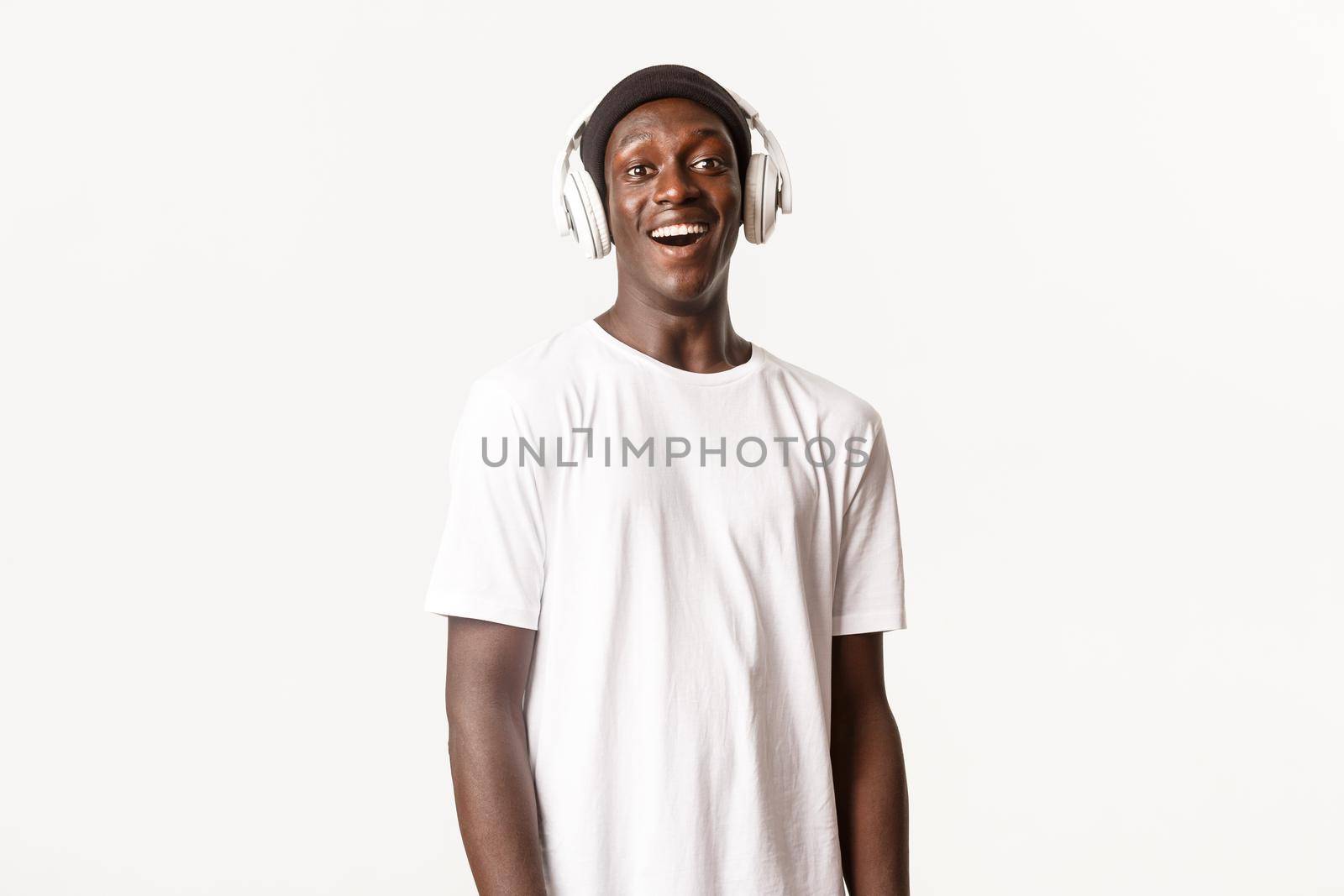 Portrait of amused and happy african-american young guy listening podcast or music in headphones, smiling pleased, white background.