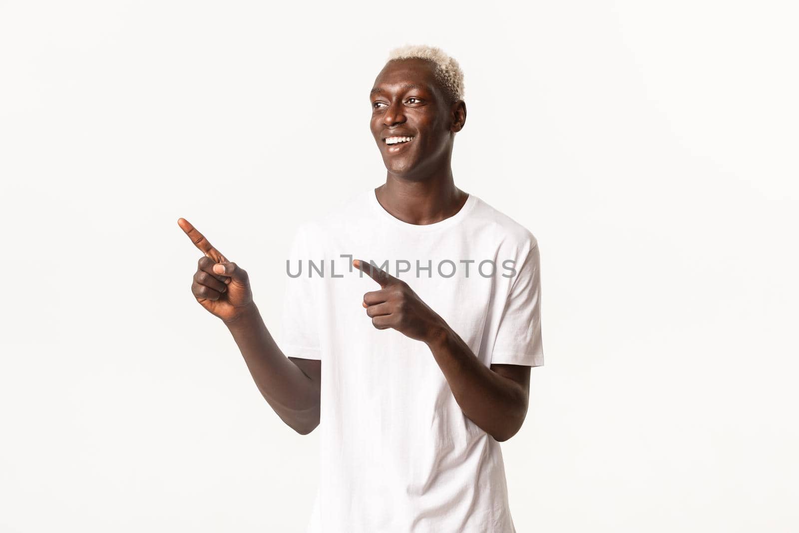 Portrait of cheerful handsome Black guy with blond hair, standing over white background, smiling pleased and pointing fingers upper left corner.