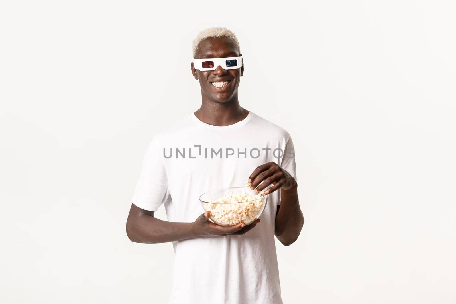 Portrait of happy attractive african-american blond guy eating popcorn and watching movie in 3d glasses, smiling cheerful, standing white background.