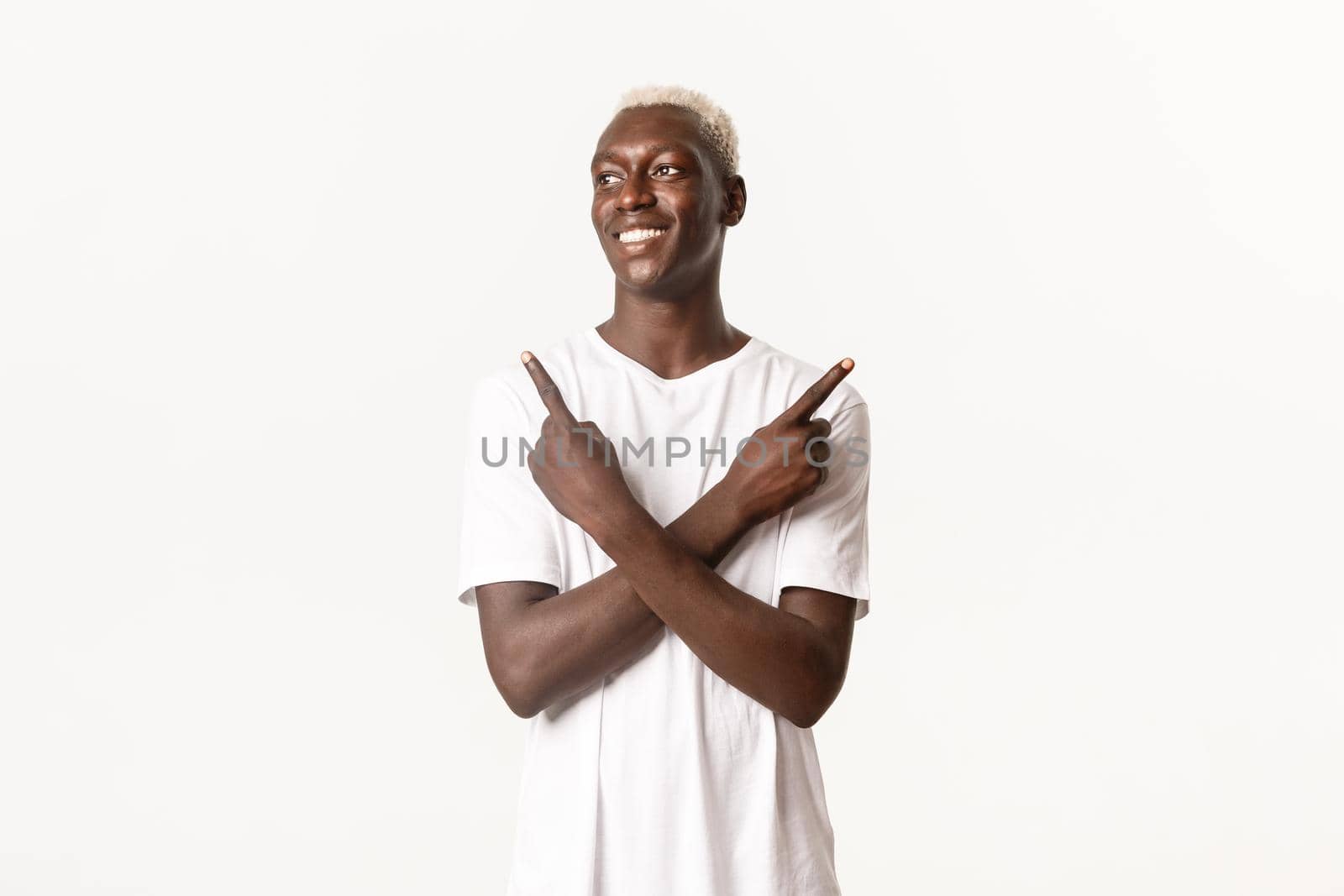 Portrait of hopeful smiling african-american guy, looking left pleased, pointing fingers sideways, standing white background.