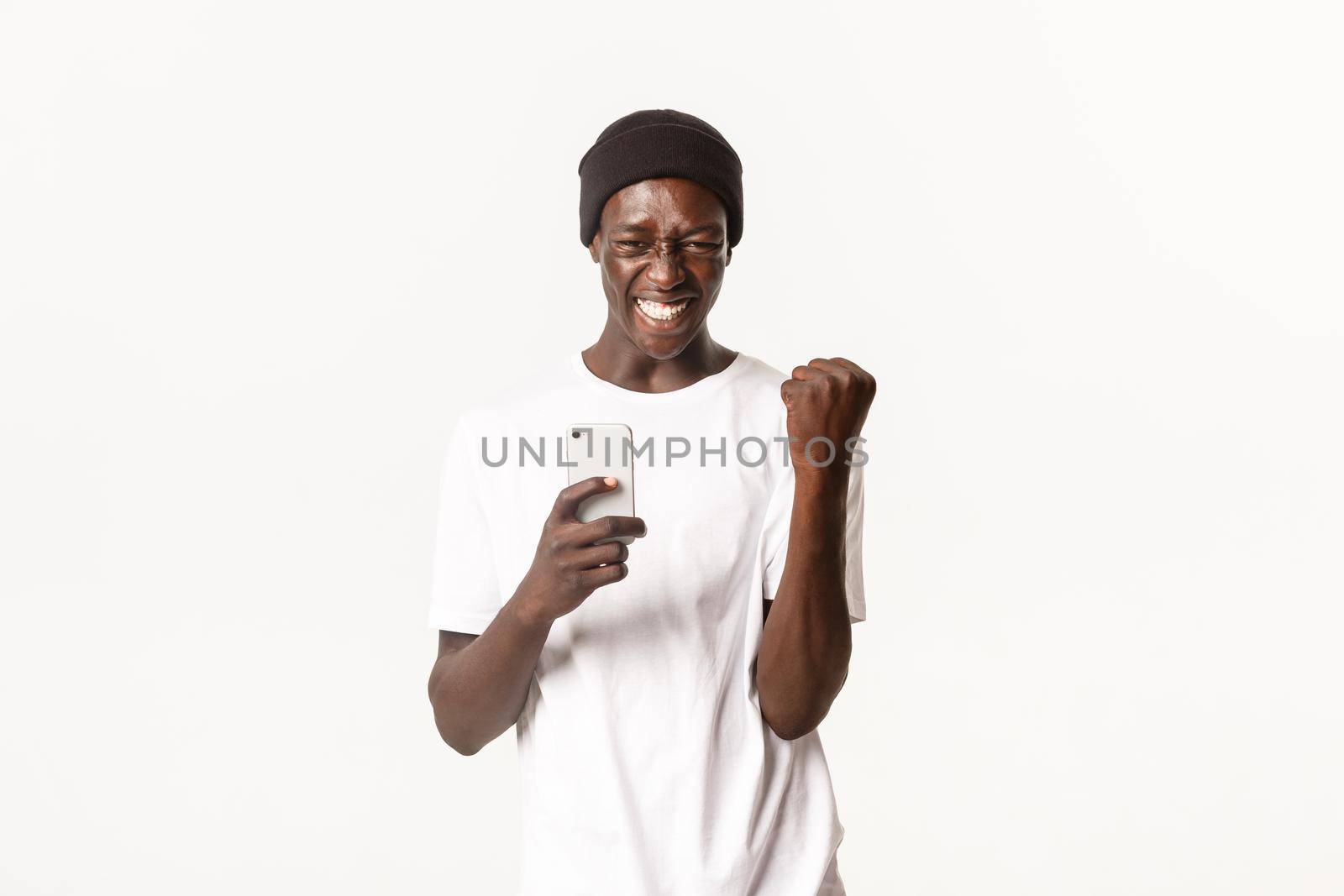 Portrait of lucky and happy african-american male in beanie, fist pump in rejoice, triumphing and smiling while using smartphone, white background.