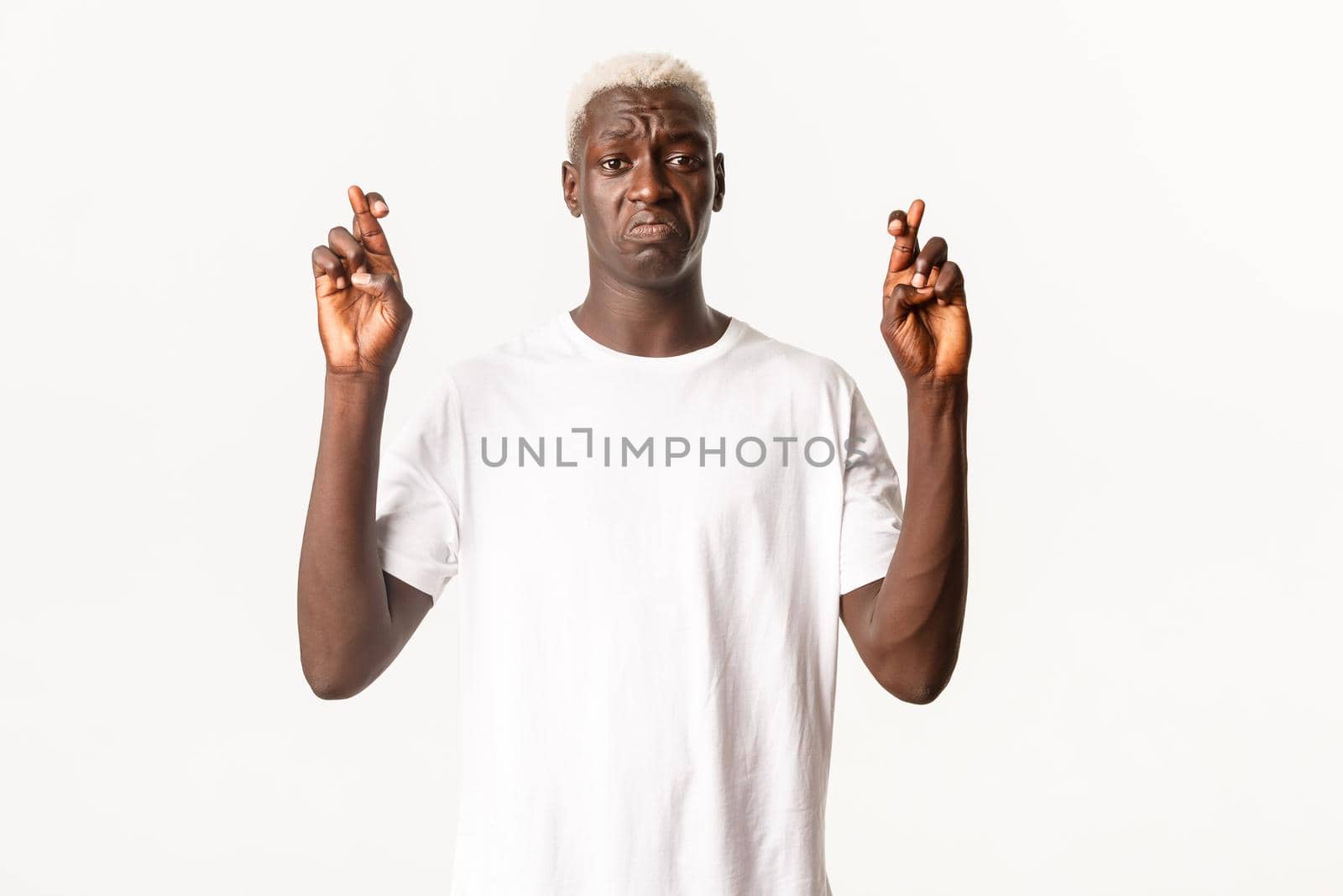 Portrait of anxious and sad african-american blond man, sulking and cross fingers good luck, making wish, white background.