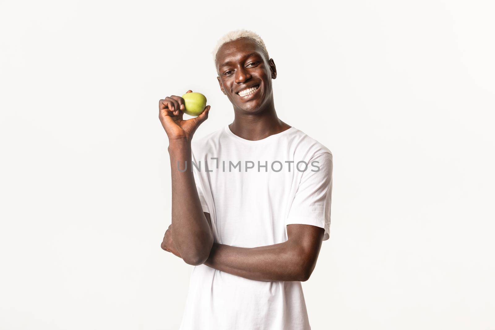 Portrait of satisfied, handsome and cheerful african-american blond man showing apple and smiling with perfect white teeth, standing in studio.