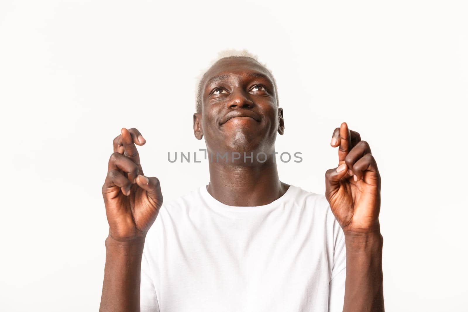 Close-up of hopeful young african-american male with blond hair, looking up sky and praying, begging god while making wish with fingers crossed, standing white background.