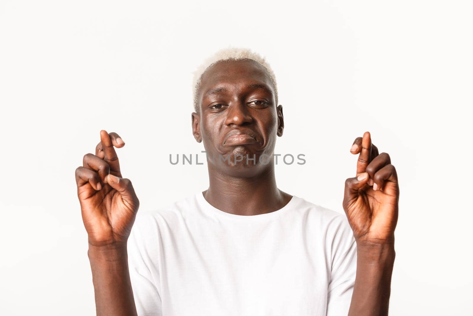 Close-up of sad and worried african-american handsome blond guy, making wish, looking upset, cross fingers good luck, praying over white background.
