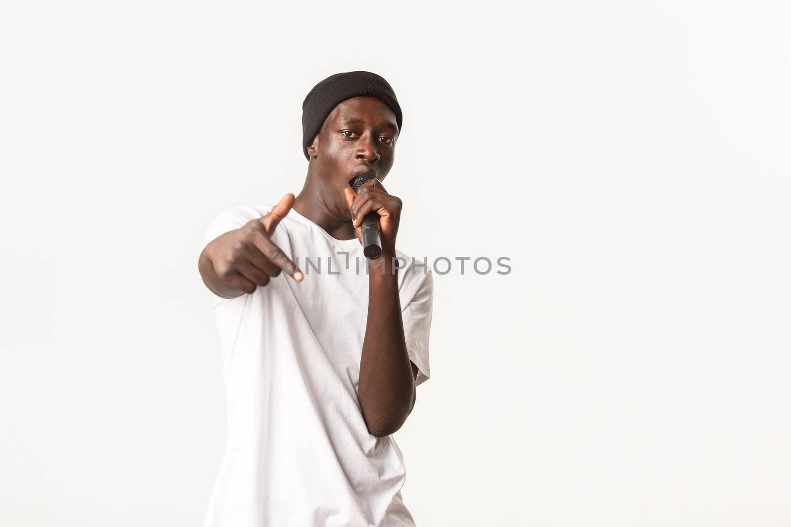 Portrait of sassy and cool Black rapper in beanie, singing into microphone and making hip-hop gesture, white background.