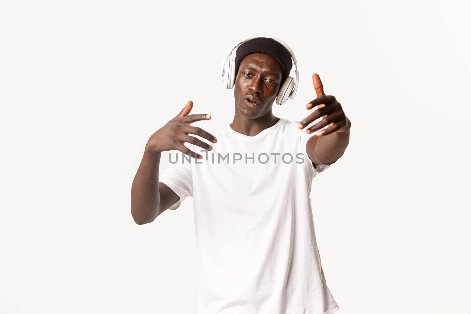 Portrait of joyful cool african-american guy, dancing and listening music in headphones, white background.
