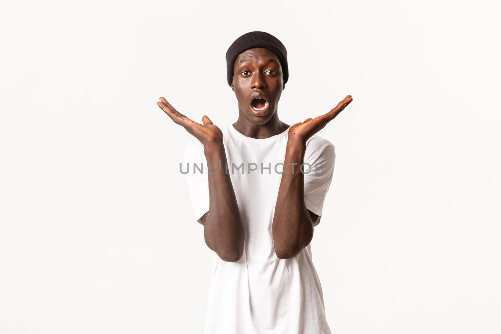 Portrait of surprised and confused handsome african-american male, open mouth and spread hands sideways amazed, standing white background in beanie and t-shirt.
