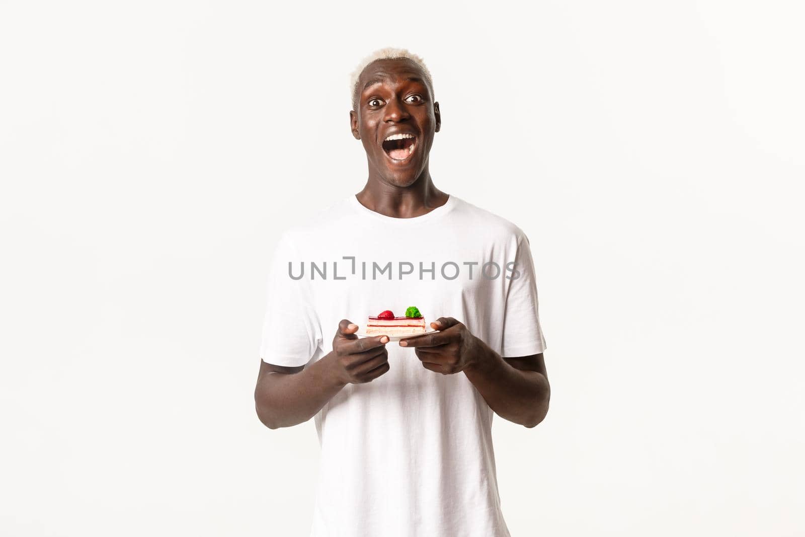 Portrait of excited, happy african-american blond guy, open mouth fascinated, holding piece of cake, standing white background.