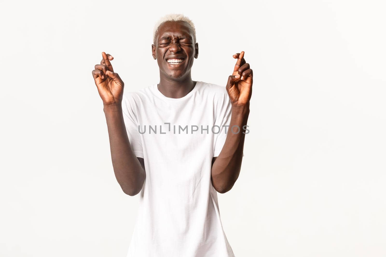 Portrait of begging hopeful african-american blond man, close eyes and cross fingers good luck, making wish, white background.