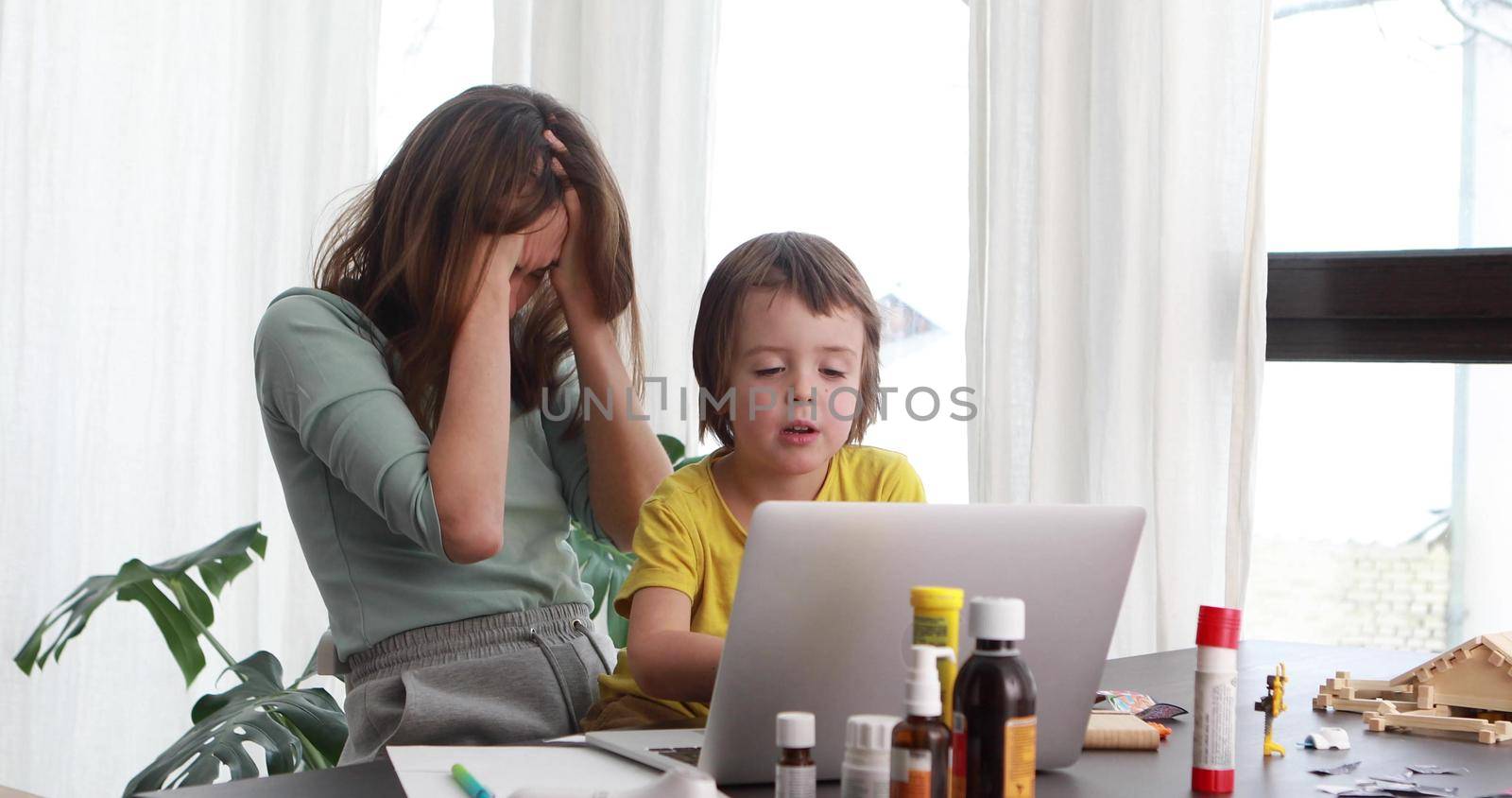 Tired woman clutches head while working at home while little boy presses keys on laptop keyboard at table with pharmacy bottles