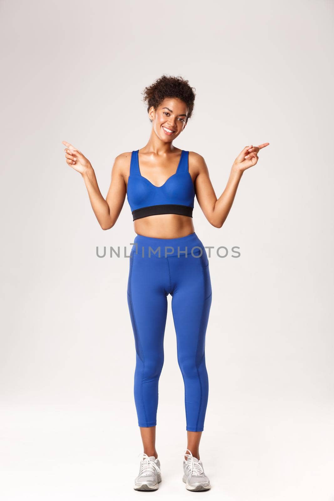 Full length of beautiful slim african-american sportswoman, wearing blue sport outfit, pointing fingers sideways, showing left and right logo, smiling happy at camera, white background.