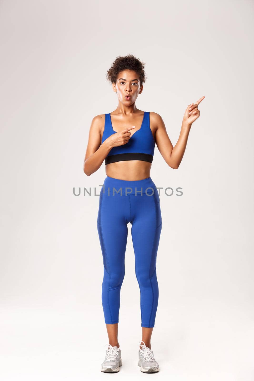 Full length of impressed and surprised african-american athletic girl, wearing sportswear, pointing fingers at upper right corner, saying wow, standing over white background.