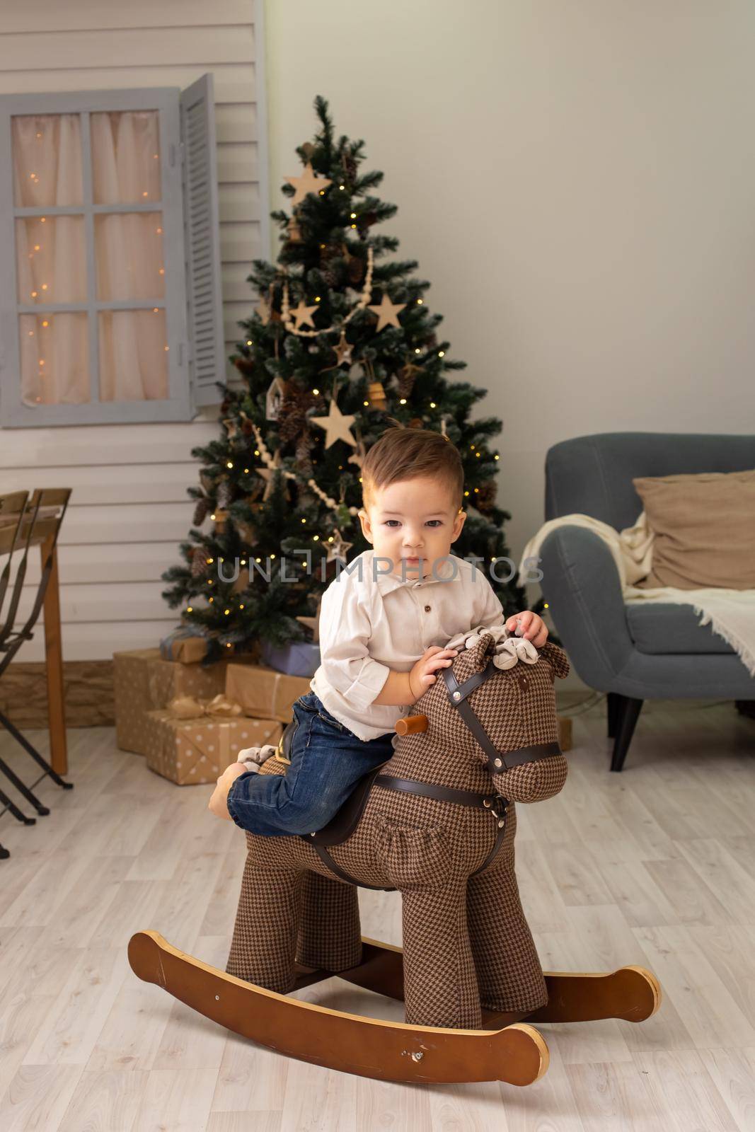Portrait of little toddler sits on a brown textile rocking horse, in the living room, with a Christmas tree by Zakharova