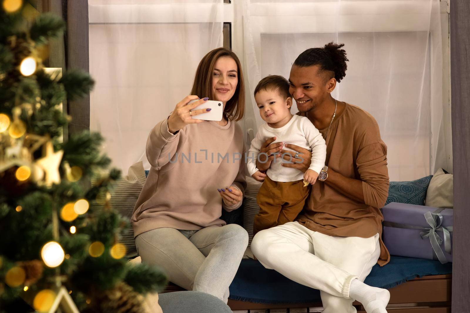 Happy beautiful young family in cozy sweatshirts: a girl with a small son, a stylish man, sitting at home, taking a photo on a smartphone and congratulating friends on the Internet Merry Christmas