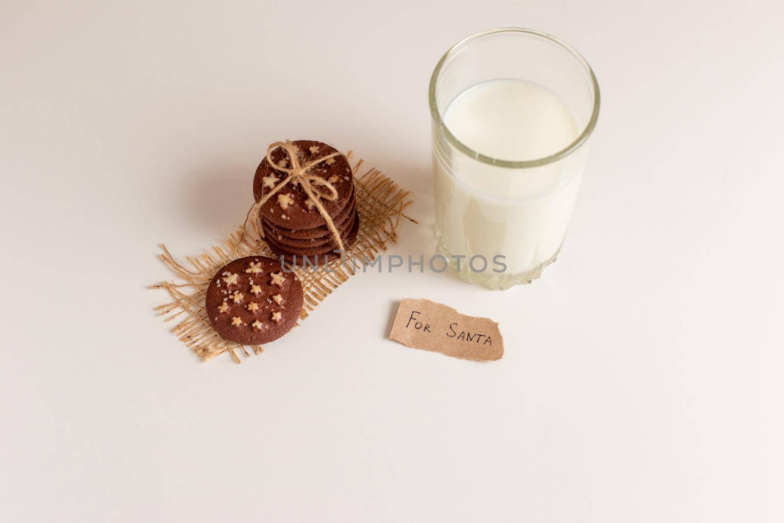 on a white table, a stack of chocolate chip cookies, a glass of milk and a small note made of paper with the inscription - for Santa. View from above