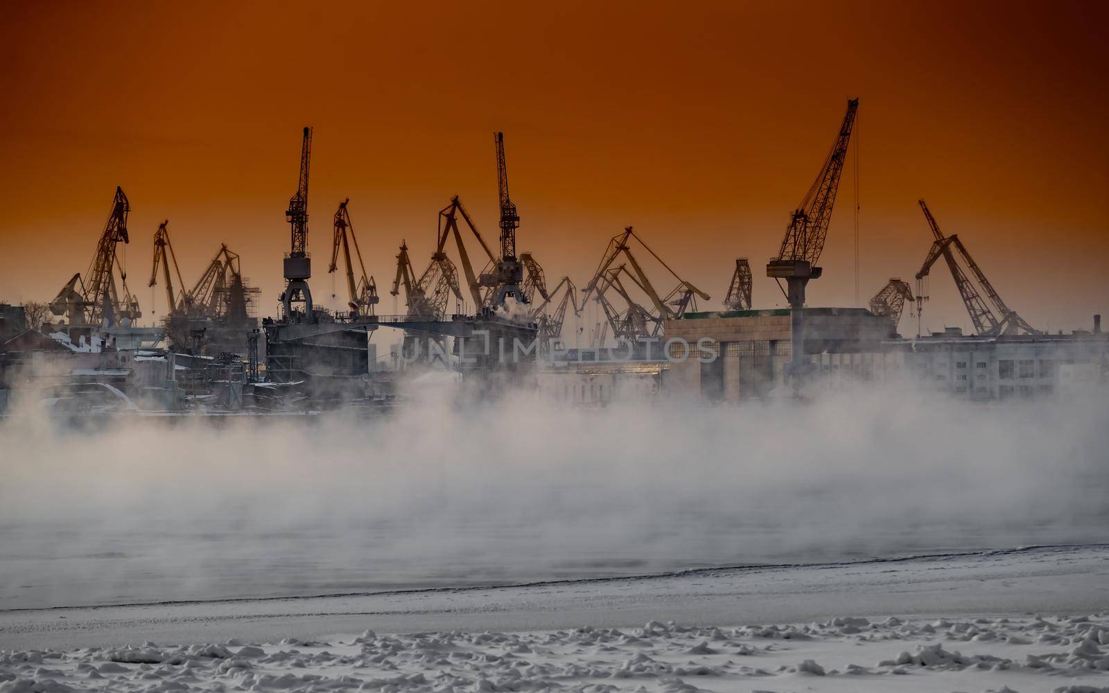 The construction of nuclear icebreakers at magic sunset, cranes of of the Baltic shipyard in a frosty winter day, steam over the Neva river, smooth surface of the river, sky of orange color. High quality photo
