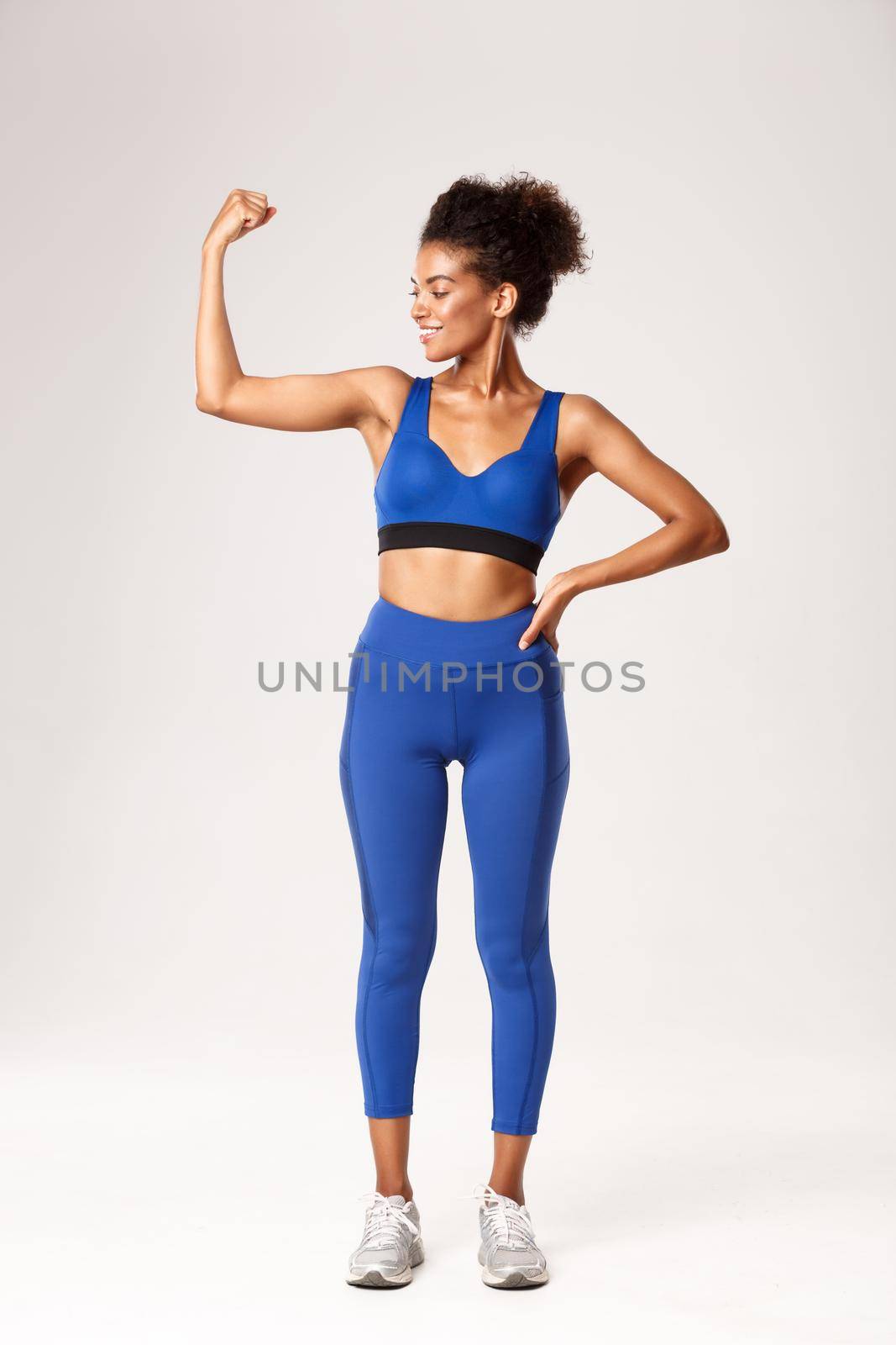 Full length of satisfied smiling african-american woman with perfect body, wearing sportswear, flex biceps and looking proud at muscles, standing over white background by Benzoix