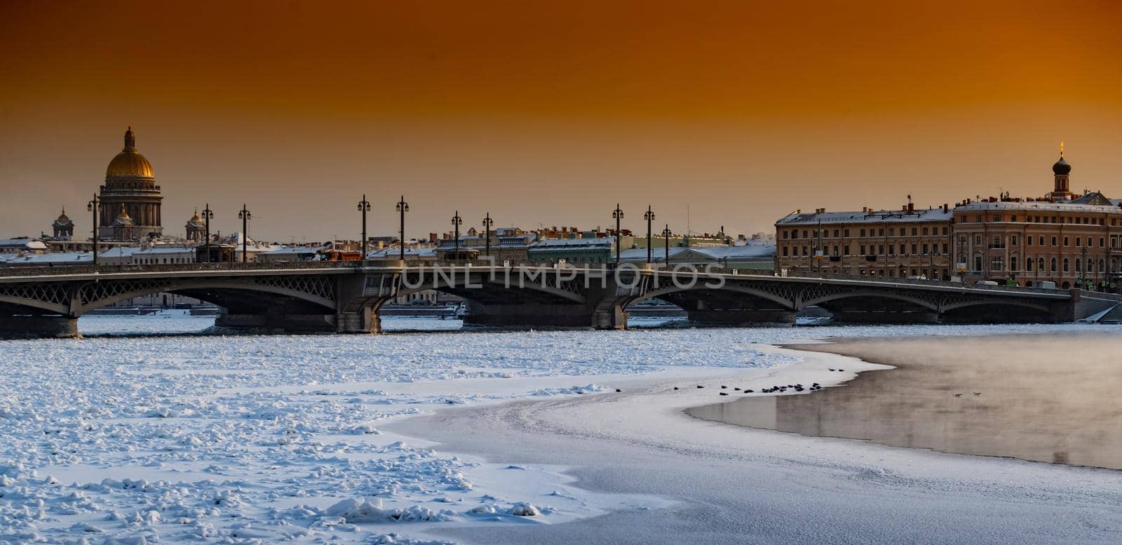 Winter panoramic view of St. Petersburg at sunset, Isaac cathedral and Blagoveshenskiy bridge on background, steam over frozen Neva river, sky of orange color. High quality photo