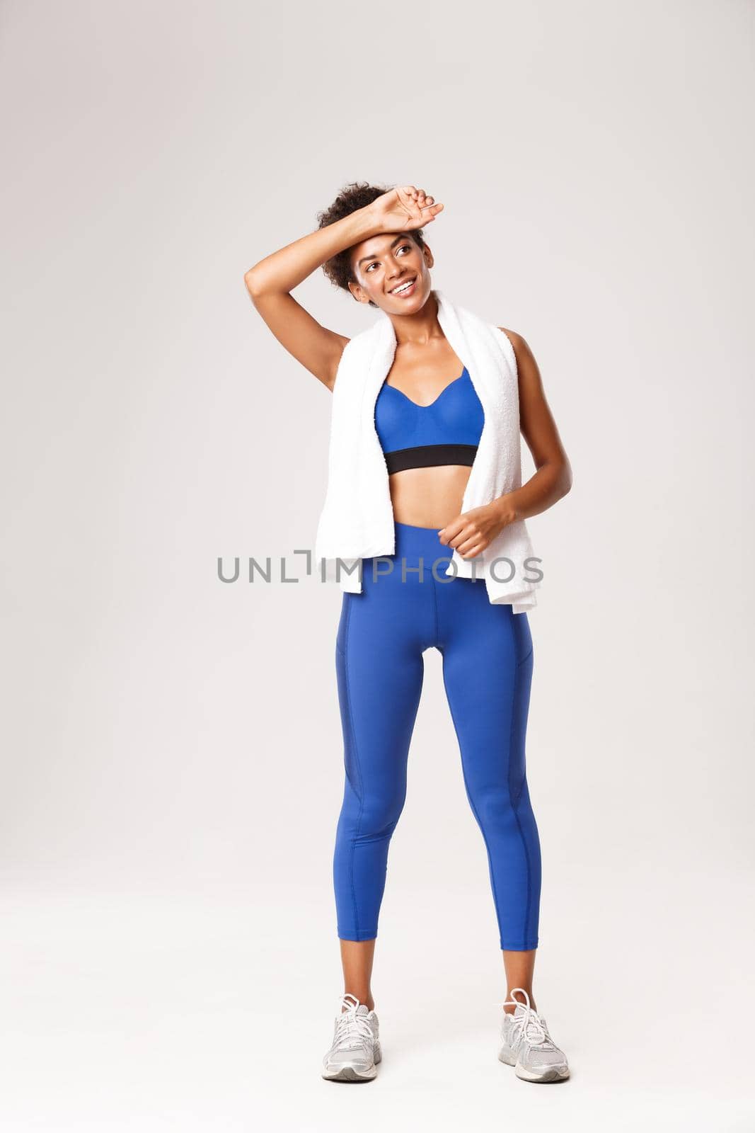 Full length of healthy and happy african-american fitness woman in blue sportswear, standing with towel after productive workout, wiping sweat off forehead with pleased smile.