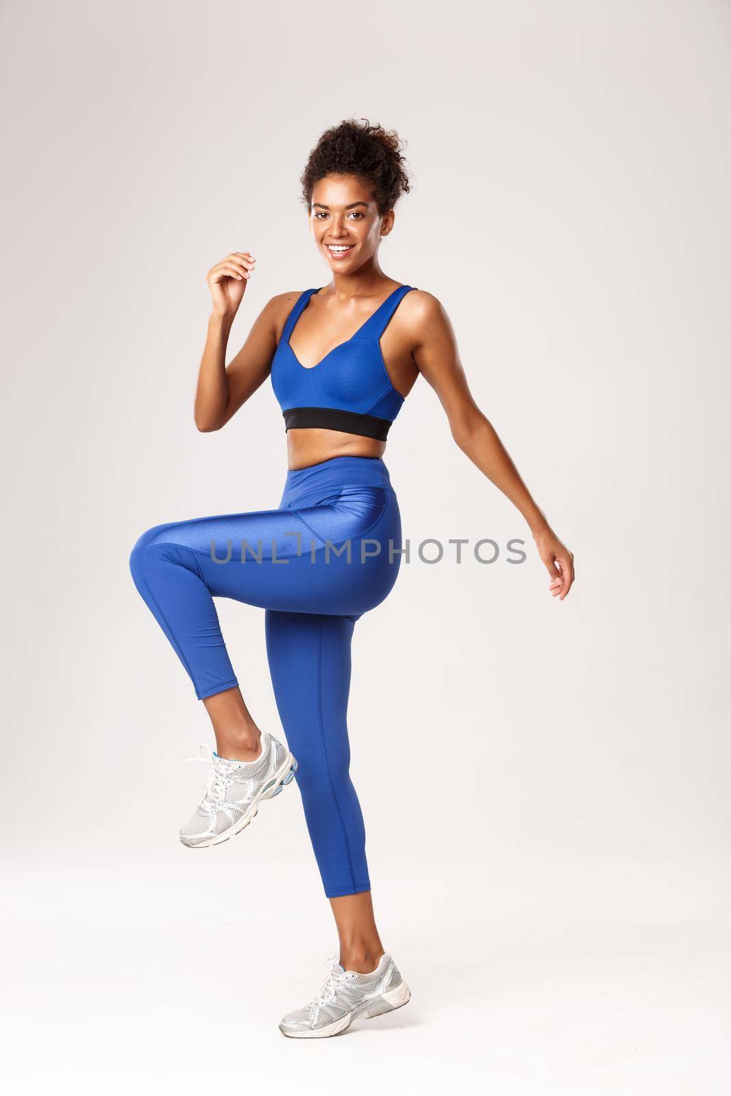 Full length of smiling african-american fitness girl workout in blue sportswear, doing leg exercises, standing over white background.