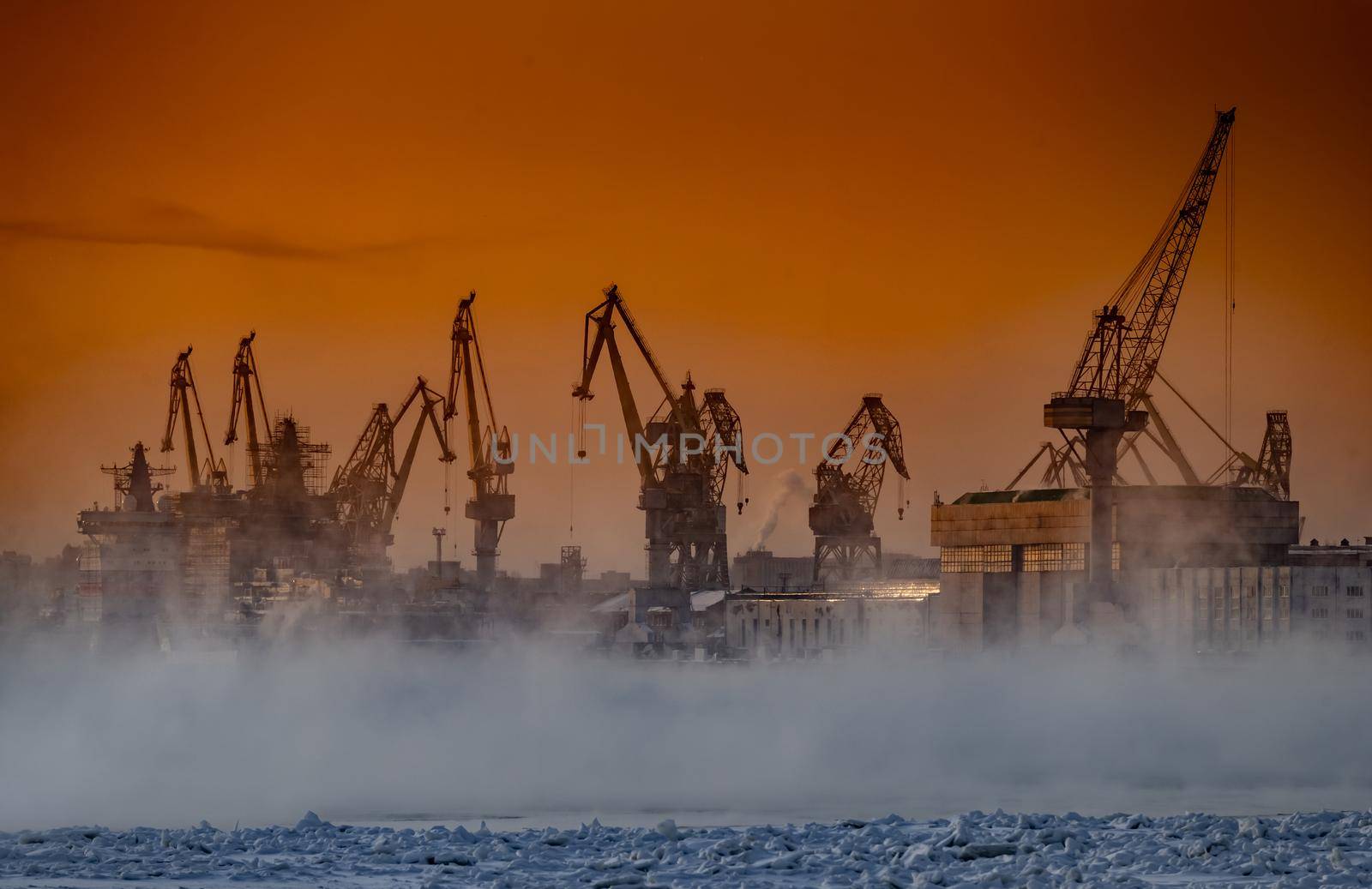 The construction of nuclear icebreakers at magic sunset, cranes of of the Baltic shipyard in a frosty winter day, steam over the Neva river, smooth surface of the river, sky of orange color. High quality photo