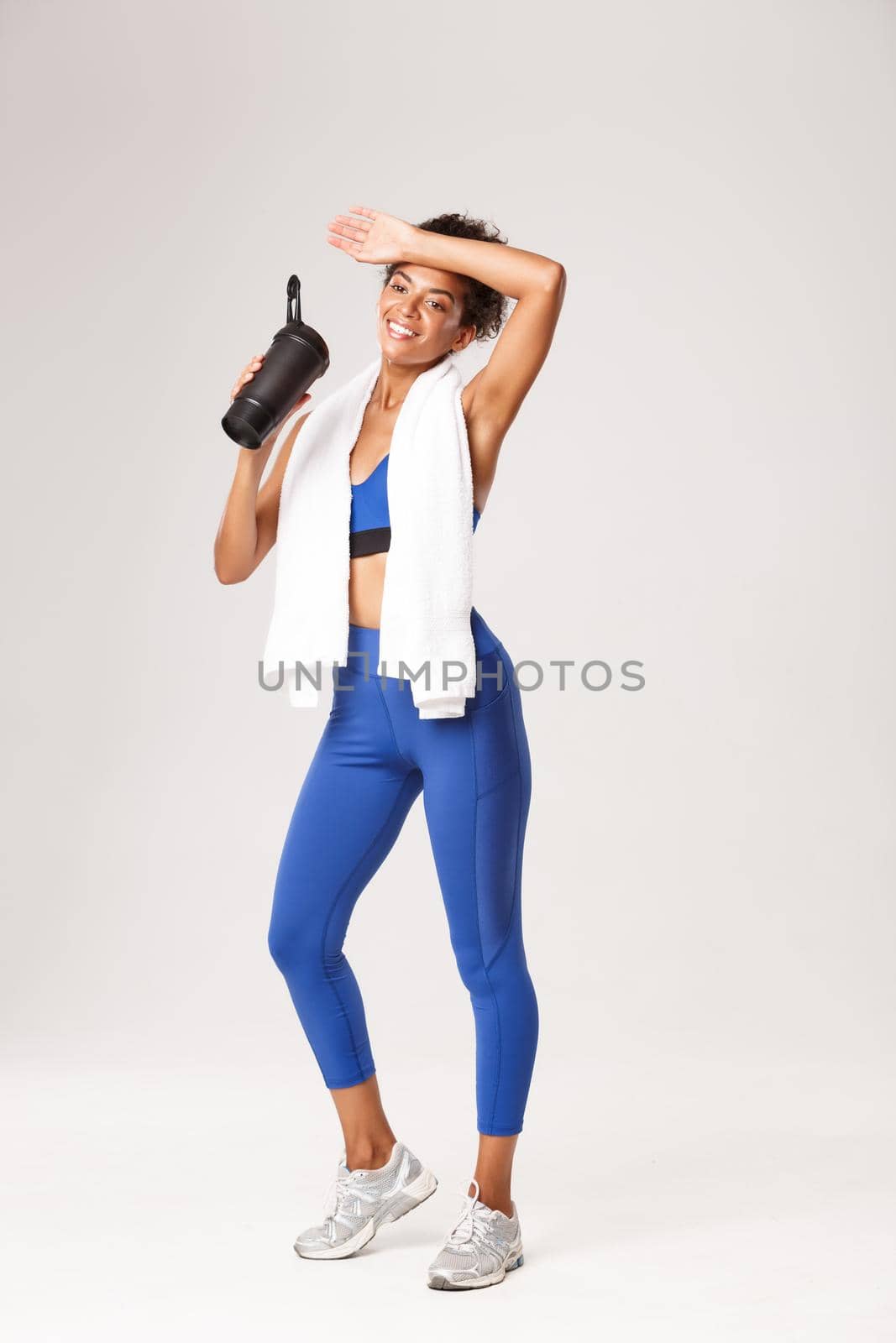 Full length of pleased smiling african-american fitness woman, drinking water, wiping sweat off forehead with satisfied face, finish good workout, standing against white background by Benzoix