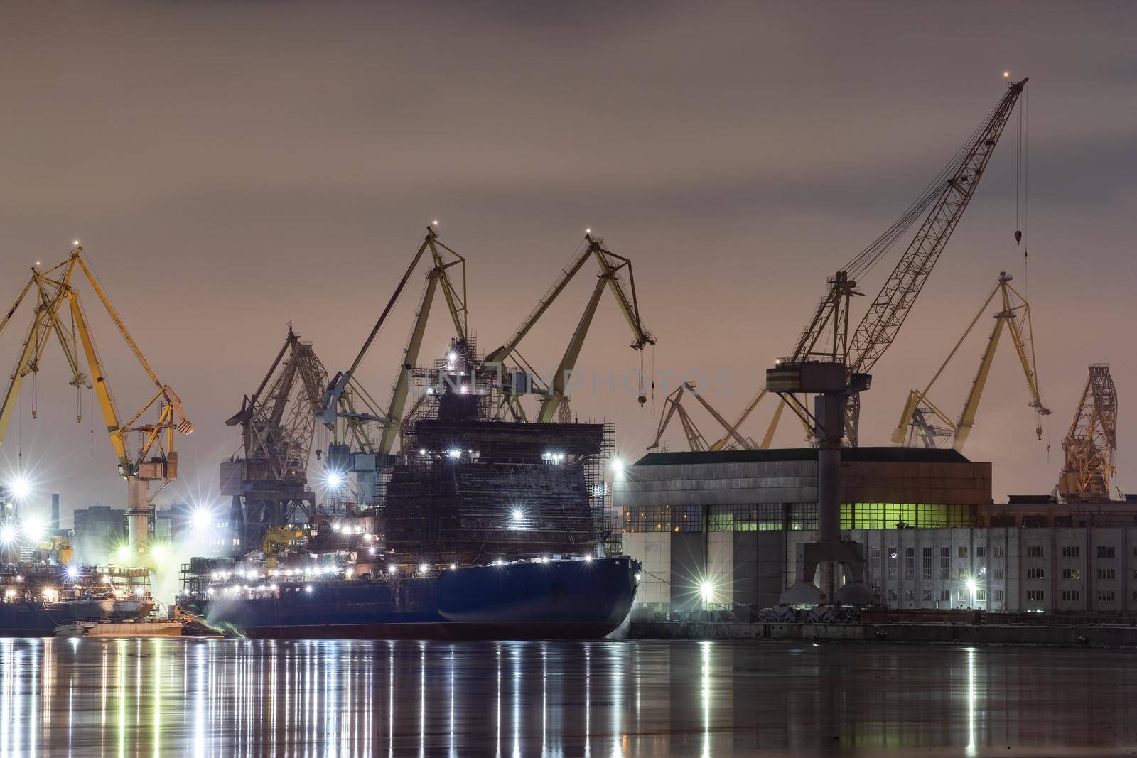 The construction of nuclear icebreakers at night, cranes of of the Baltic shipyard in a frosty winter day, steam over the Neva river, smooth surface of the river, fog. High quality photo