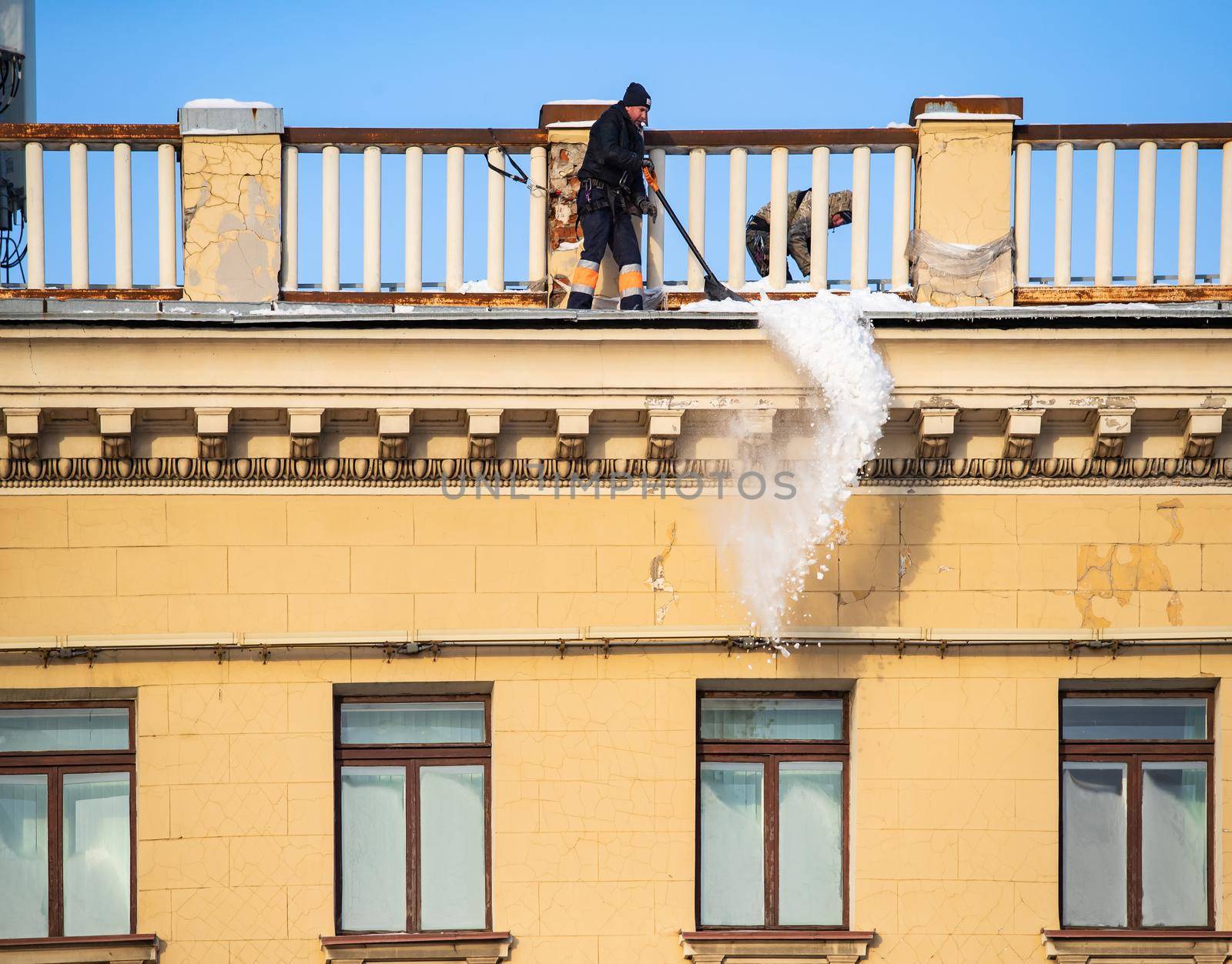 Russia, St. Petersburg, 07 December 2021: The work of utilities to remove snow from the roofs of houses, a man throws off the snow of the roof, he is attached with insurance to the roof structure. High quality photo