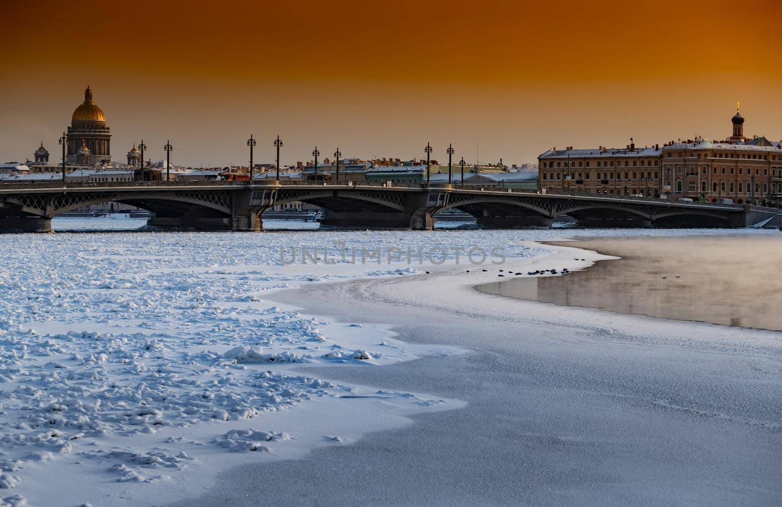 Winter panoramic view of St. Petersburg at sunset, Isaac cathedral and Blagoveshenskiy bridge on background, steam over frozen Neva river, sky of orange color. High quality photo