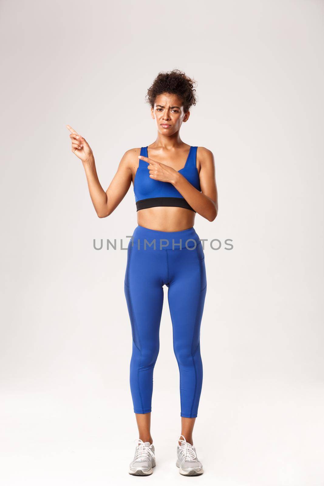 Full length of disappointed african-american girl in sportswear, looking upset, frowning and pointing fingers left at brand name, standing over white background in workout outfit.