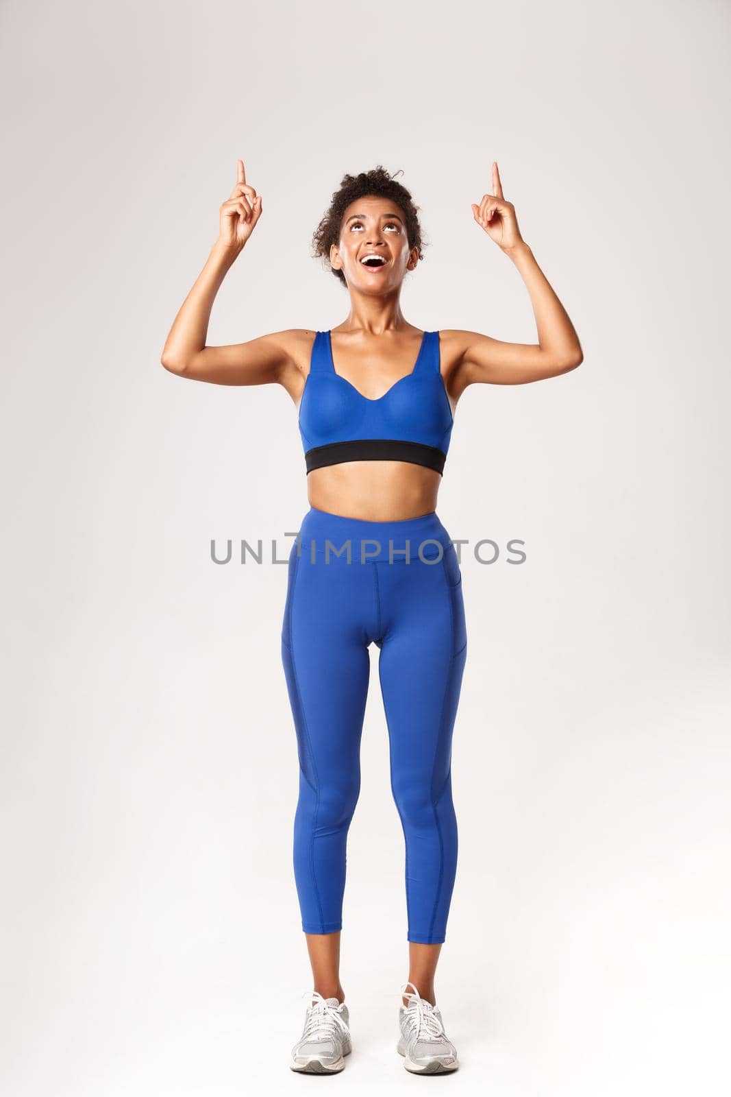 Full length of excited and happy african-american woman in sportswear for workout, looking and pointing fingers up with amazed face, white background by Benzoix