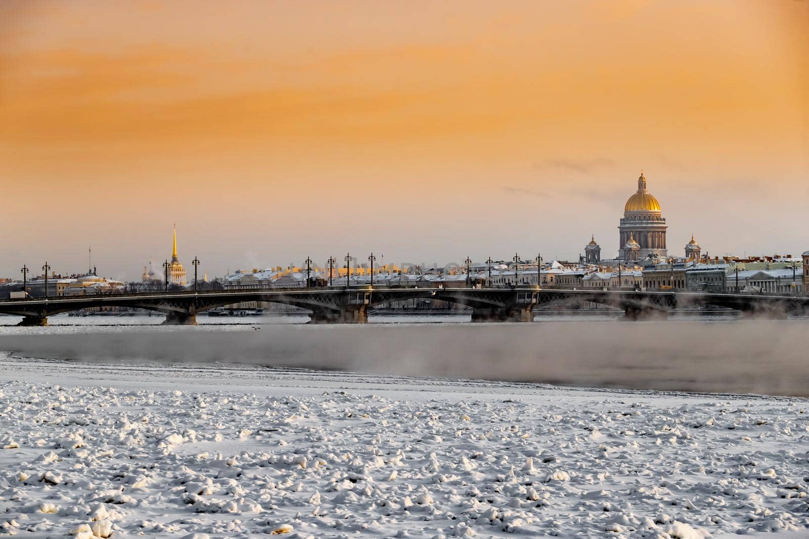 Winter panoramic view of St. Petersburg at sunset, Isaac cathedral and Blagoveshenskiy bridge on background, steam over frozen Neva river, sky of orange color. High quality photo