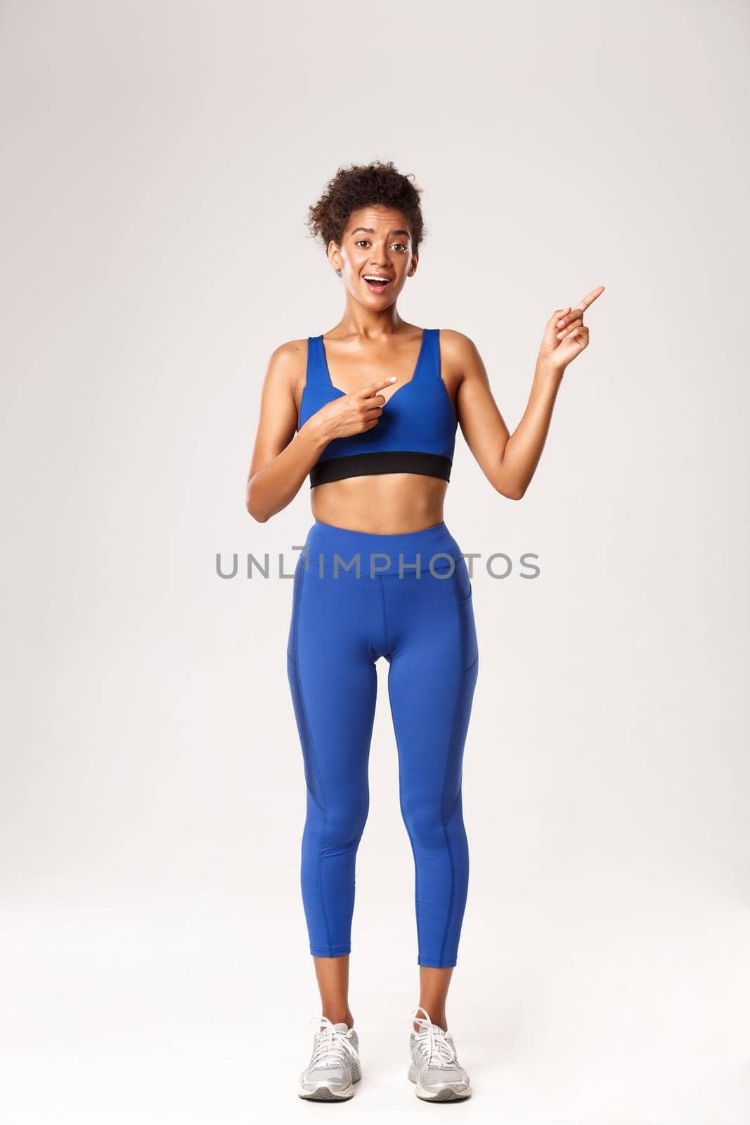 Full length of excited good-looking african-american sportswoman in fitness clothing, pointing fingers at upper right corner, standing over white background.