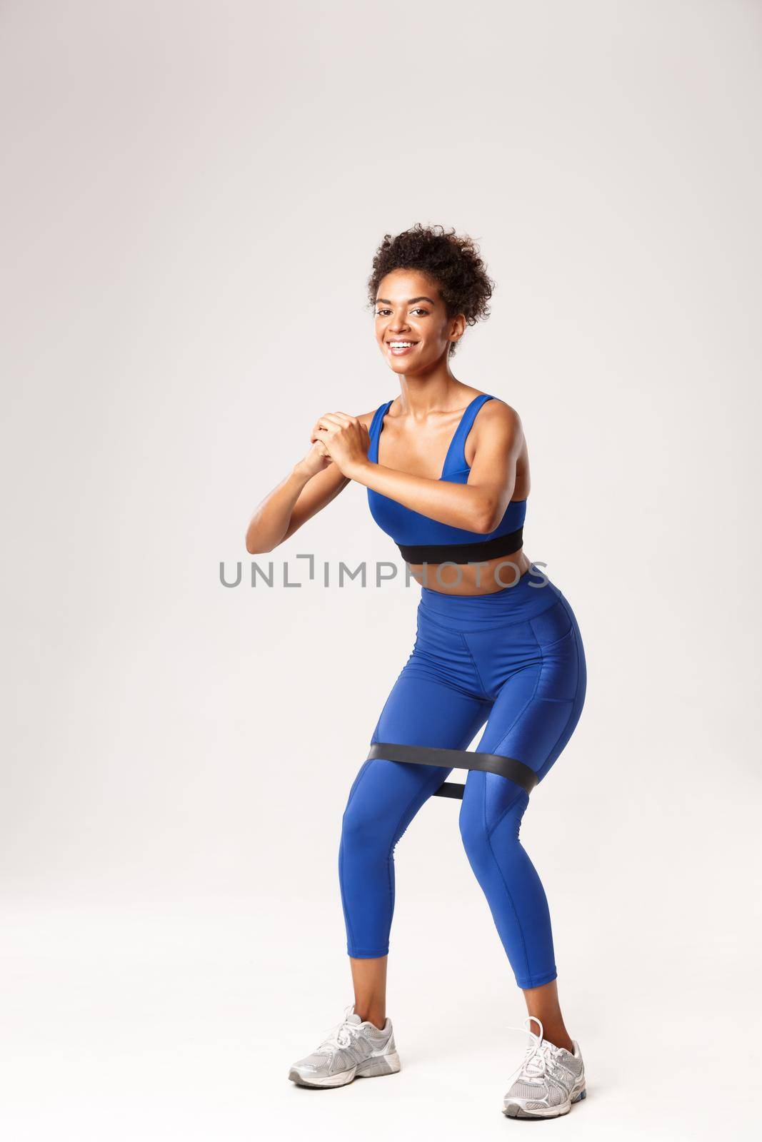 Full length of smiling fit woman enjoying fitness, wearing blue sport outfit, doing squats with resistance band, standing against white background.