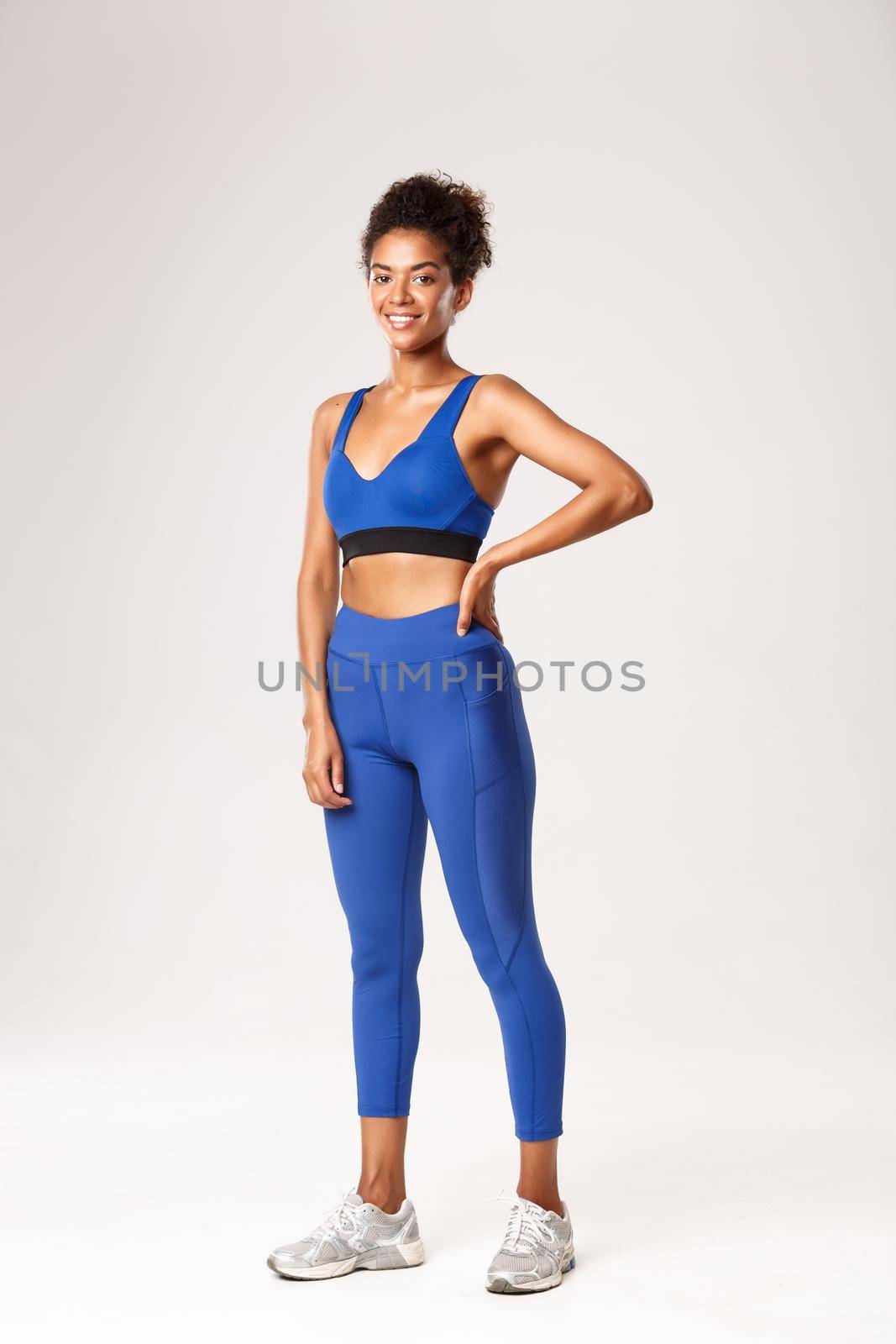 Full length of attractive african-american sportswoman with curly combed hair, wearing sports bra and leggings, smiling at camera, ready for workout, standing over white background.