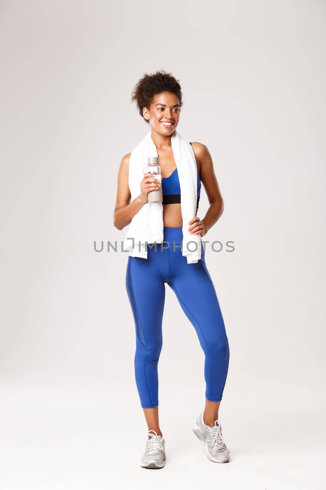 Full length of happy and healthy african-american fitness woman, wearing blue outfit, standing with water bottle and towel after exercises, posing over white background by Benzoix