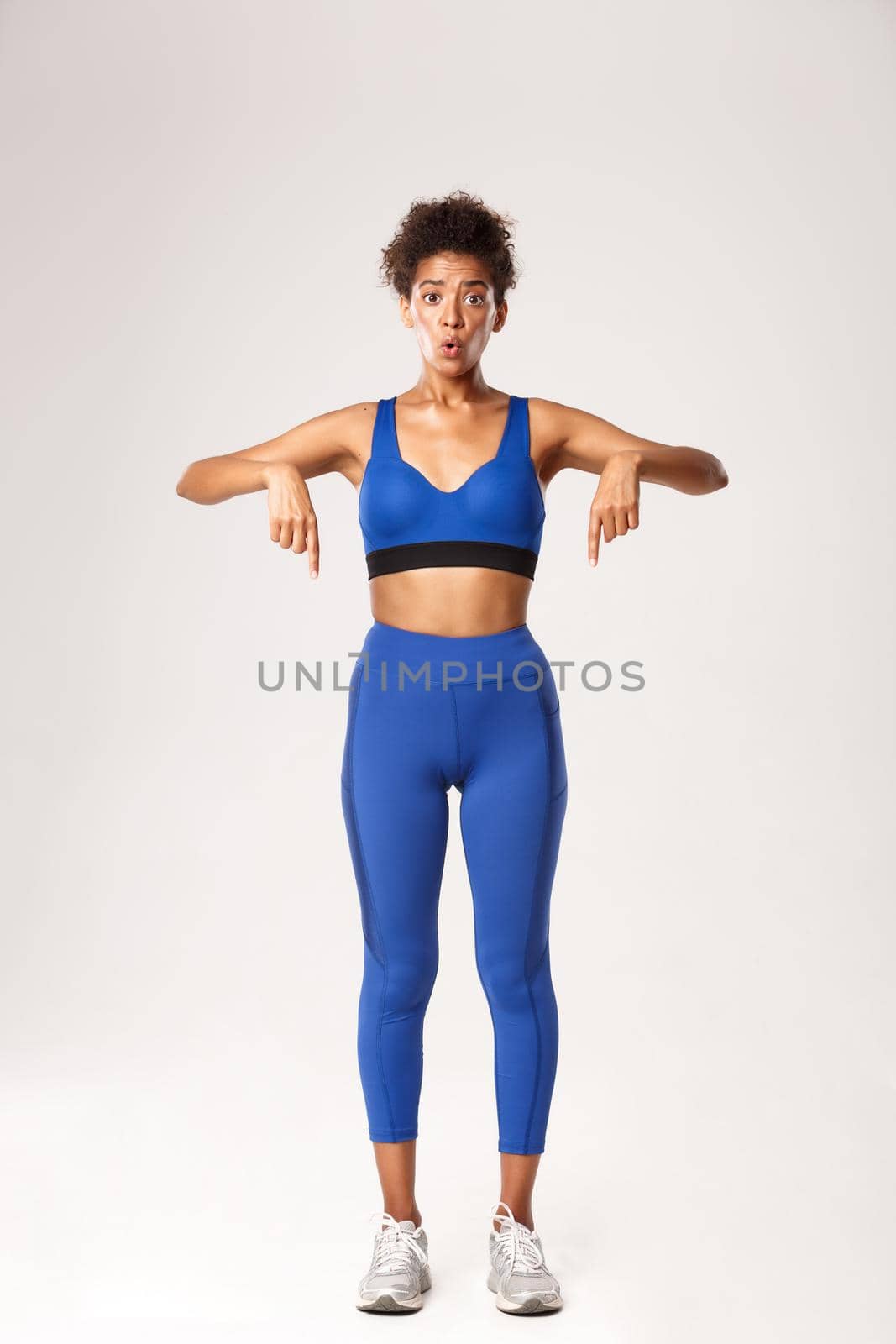 Full length of pretty african-american sportswoman looking wondered, pointing fingers down, wearing blue sportswear, white background.