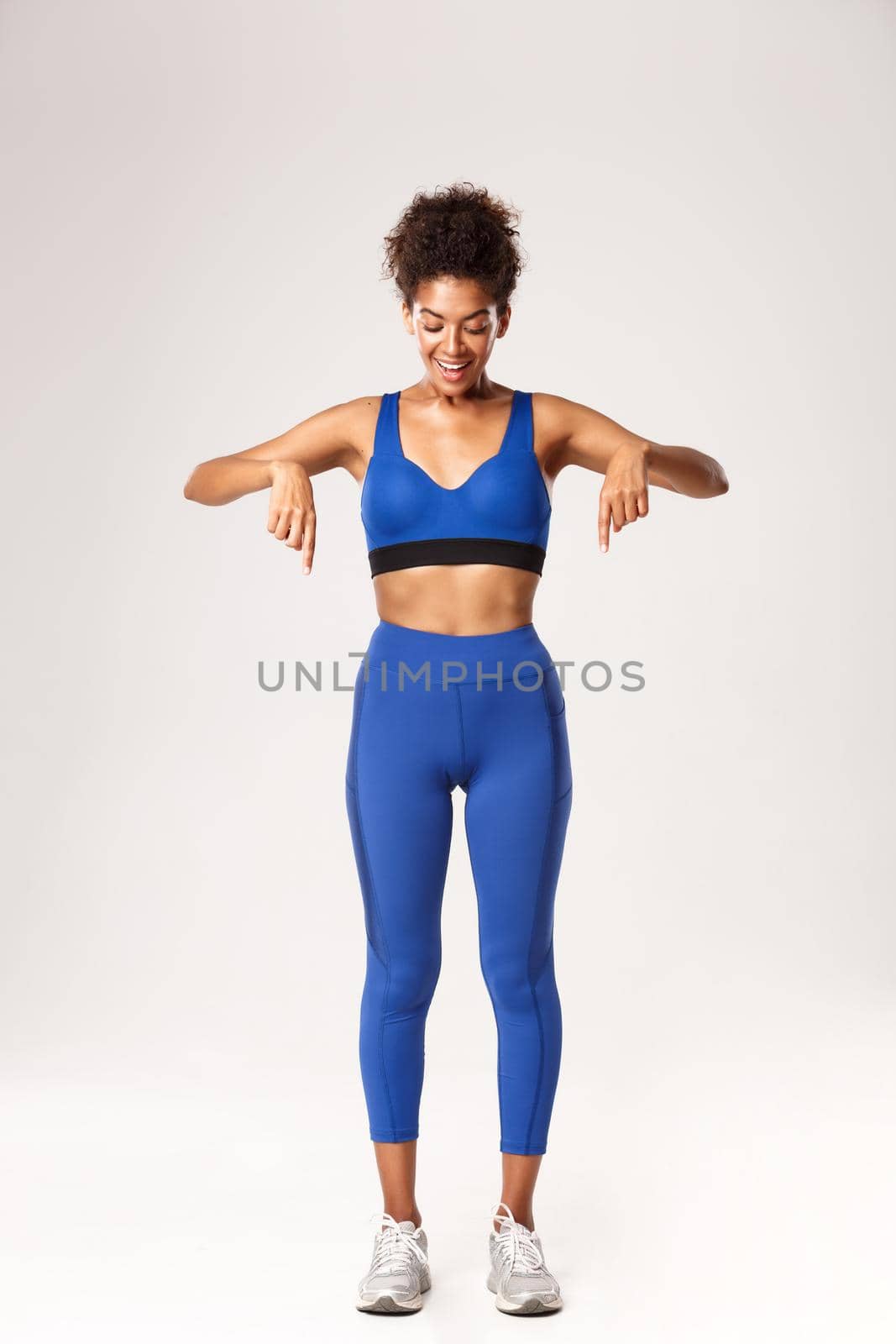 Full length of amazed african-american female athlete, wearing blue sports outfit, pointing fingers down and looking with excited smile, standing over white background by Benzoix