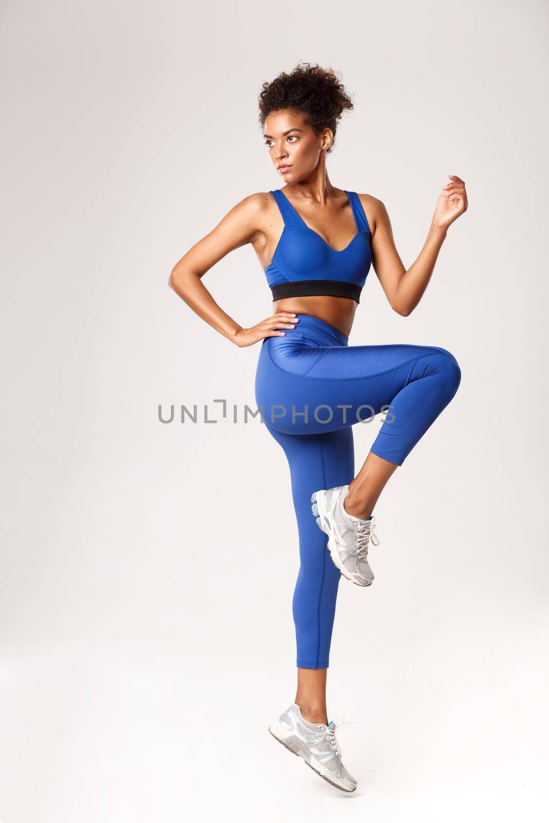 Full length of serious african-american sportswoman in blue leggings and sports bra, raising leg up, workout in gym, standing over white background by Benzoix