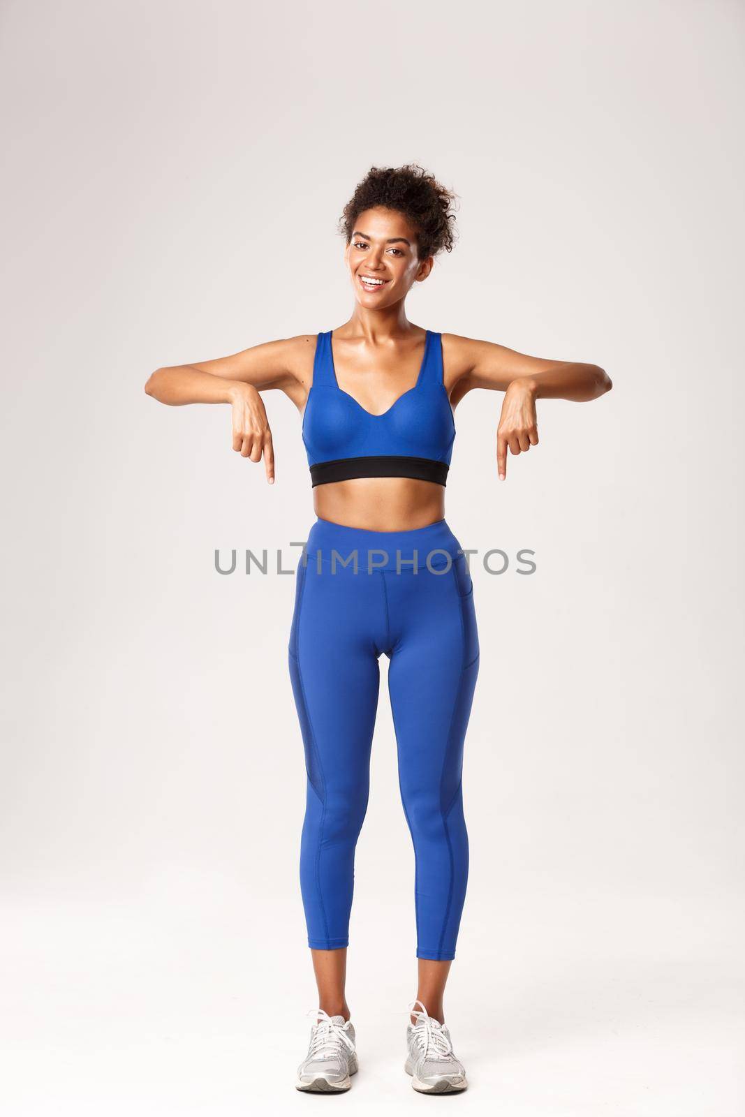 Full length of happy african-american fitness coach in blue sportswear, pointing fingers down and smiling, recommend workout gear, showing logo or sports promo, white background.