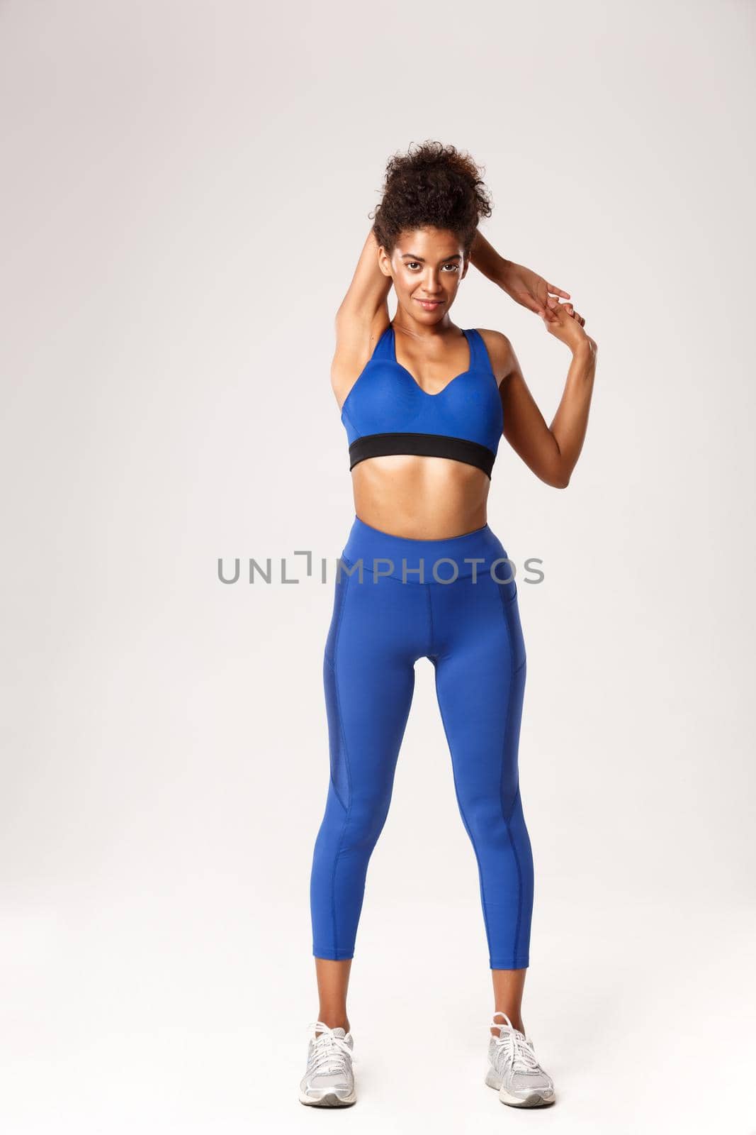 Full length of smiling attractive african-american fitness girl in blue sport outfit, stretching hands, warming-up before workout, standing against white background.