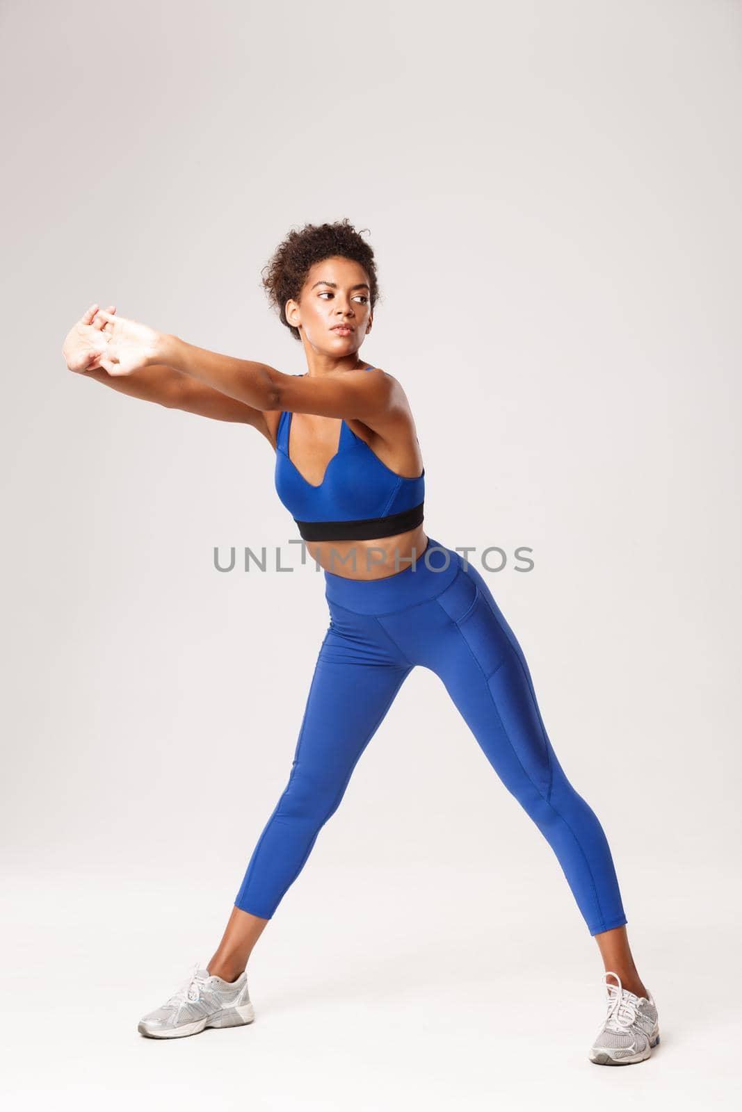 Full length of attractive african-american woman with fit body, wearing blue sportwear, exercising over white background, showing stretching warm-up workout, looking aside.