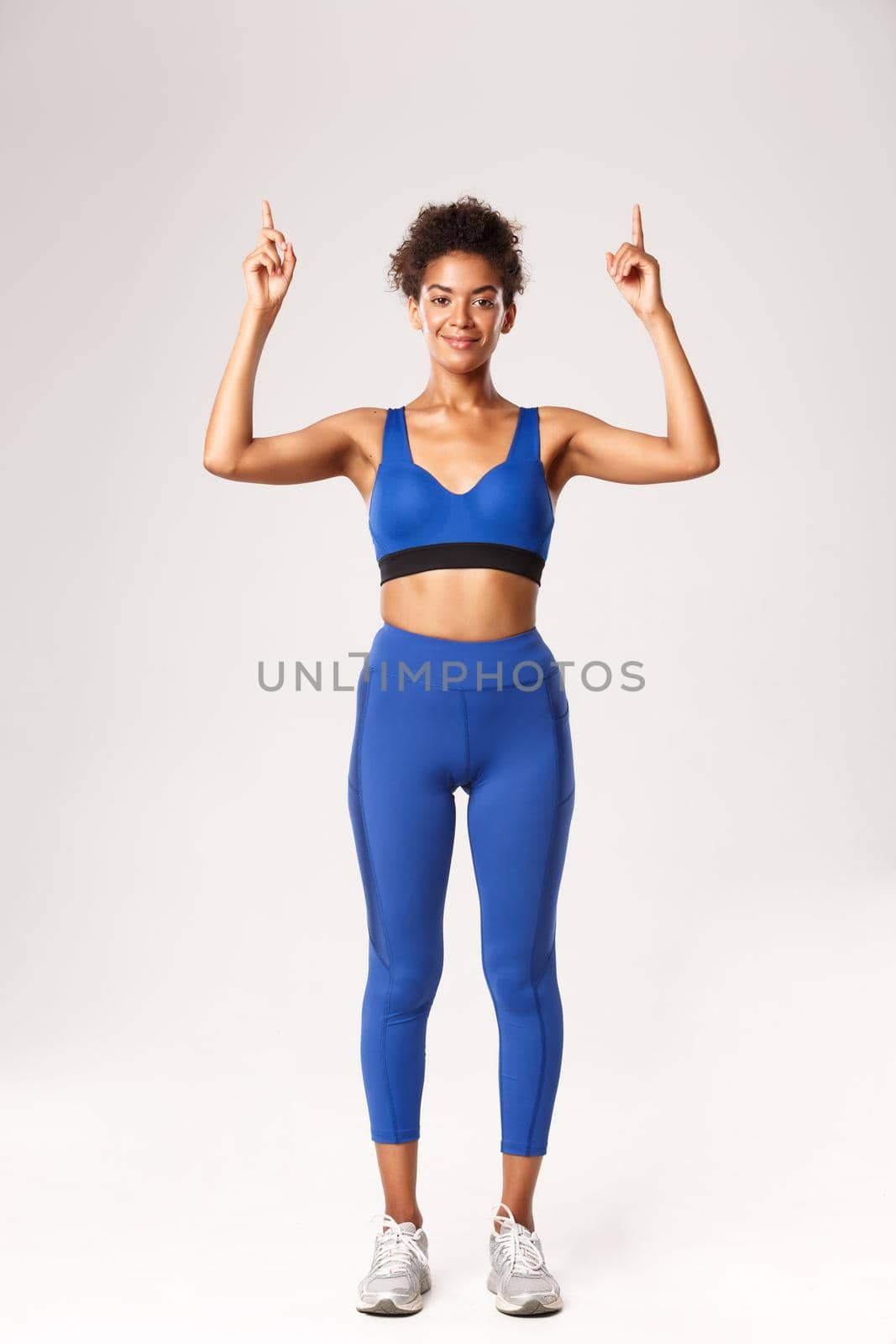 Full length of smiling attractive Black sportswoman in blue sports clothing, looking pleased at camera and pointing fingers up, showing logo or advertisement about workout, white background.
