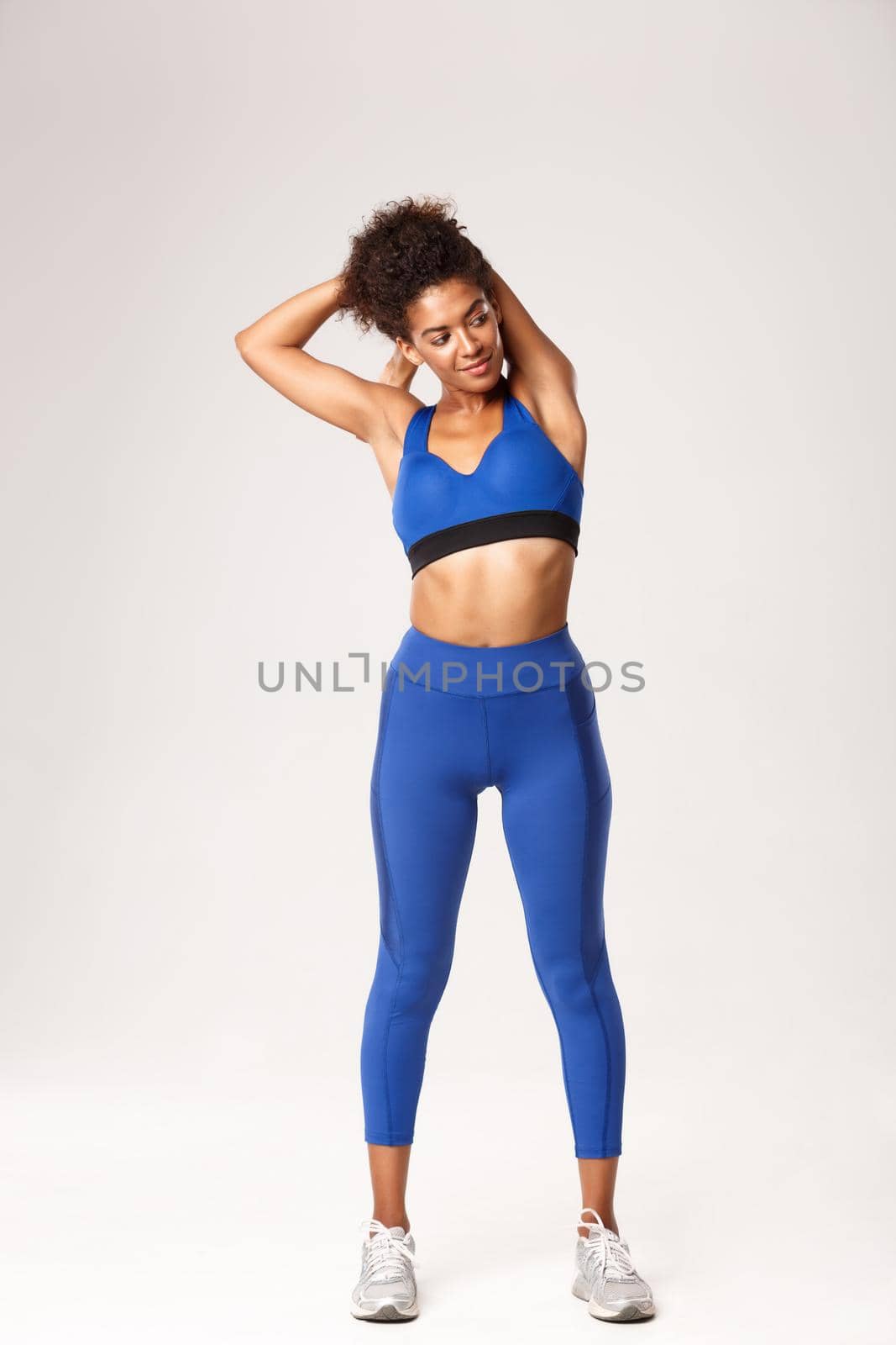 Full length of beautiful smiling african-american sportswoman, wearing blue sport outfit, warming-up before workout, showing stretching exercises, white background.