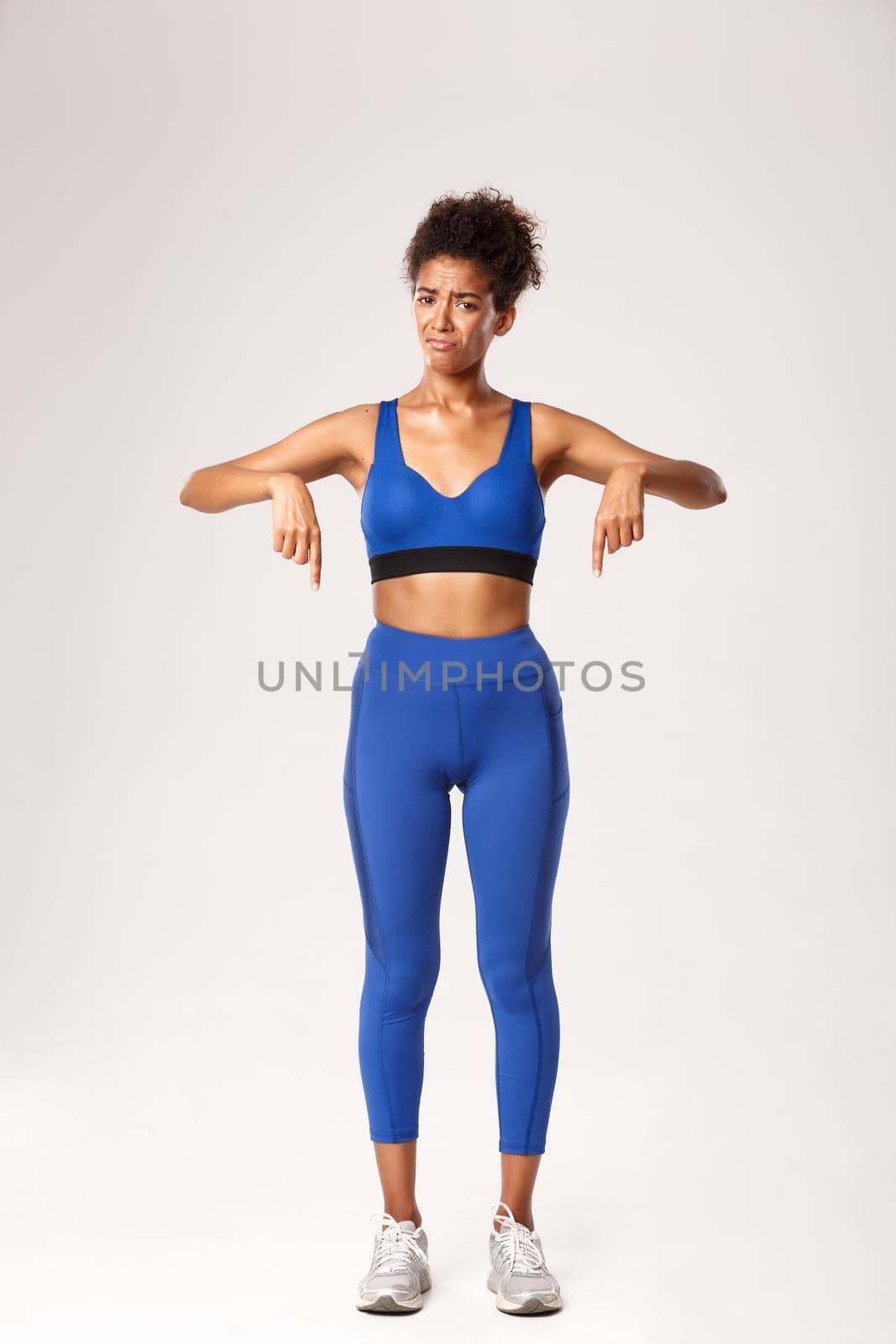 Full length of disappointed african-american girl in blue sports outfit, pointing fingers down and grimacing, complaining about something bad, standing over white background.